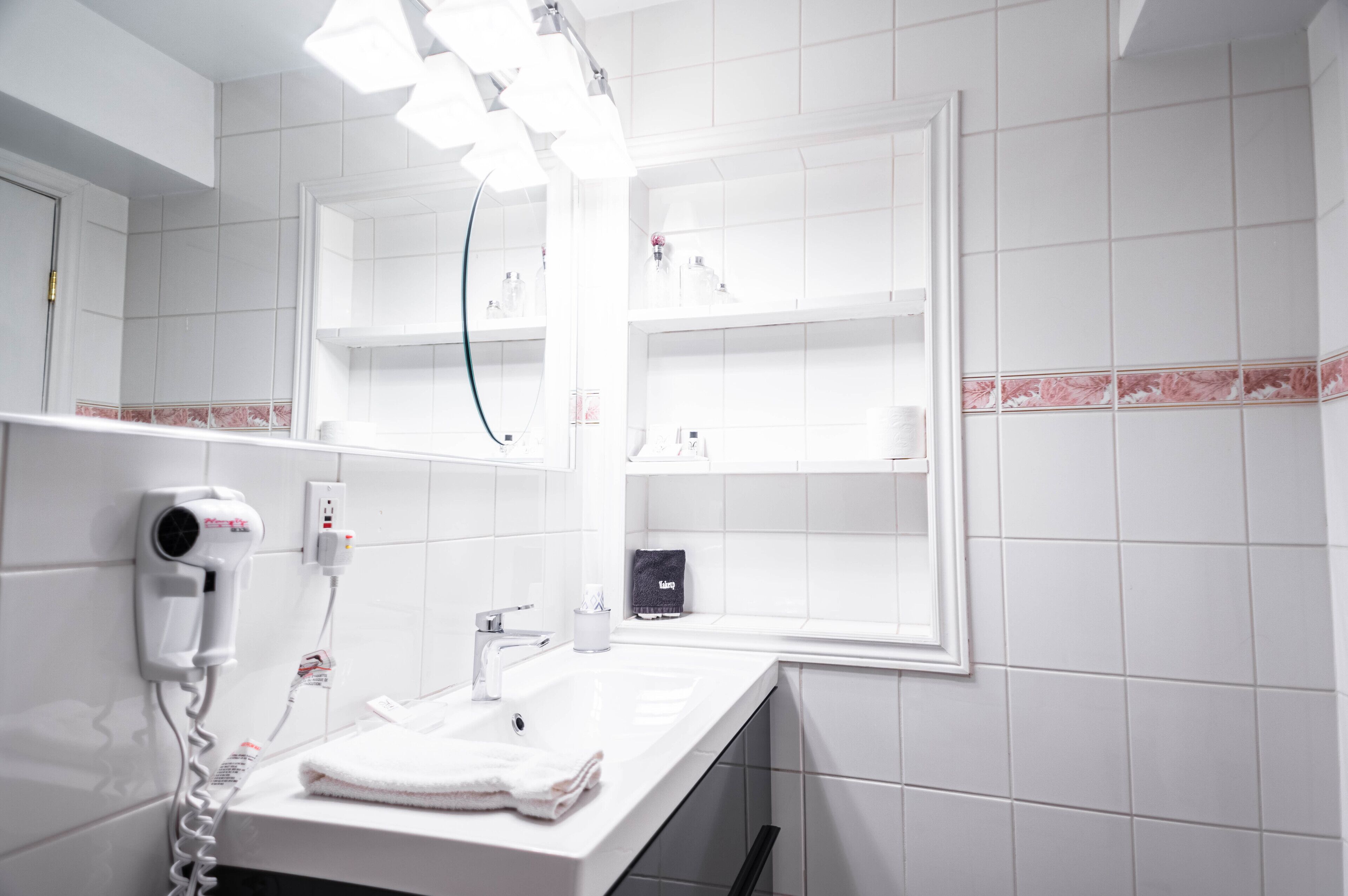 Casa de banho com lavatório branco, secador de cabelo e estantes com produtos de higiene pessoal.