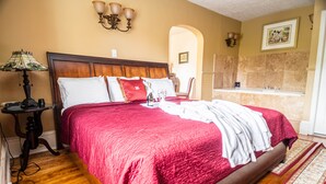 Una cama bien hecha con colcha y almohadas rojas, un cabecero de madera, una mesita de noche con lámpara, y un baño con bañera y ducha.