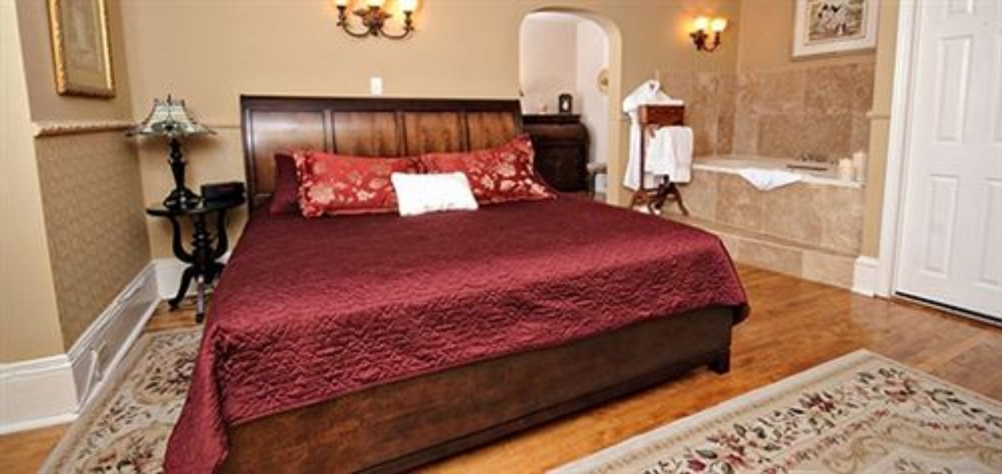Quarto com cama grande, mesa de cabeceira, abajur e banheiro visível por um arco.