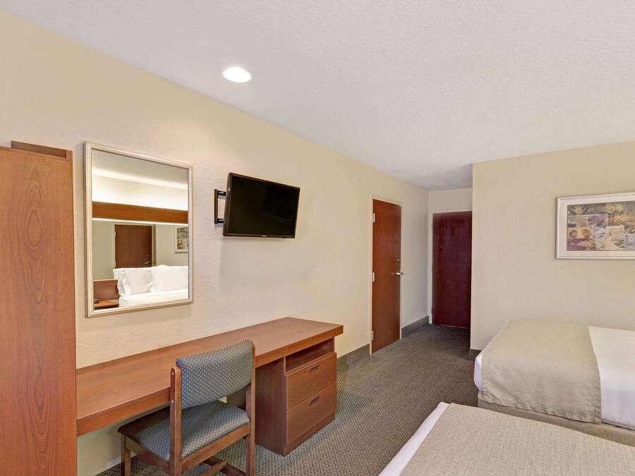A hotel room with a wooden desk, a chair, a bed, a television mounted on the wall, and a door.