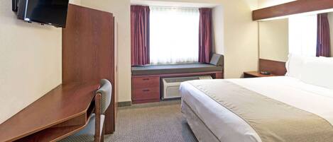 A hotel room with a bed, a desk, a chair, a TV mounted on the wall, and a window with curtains.