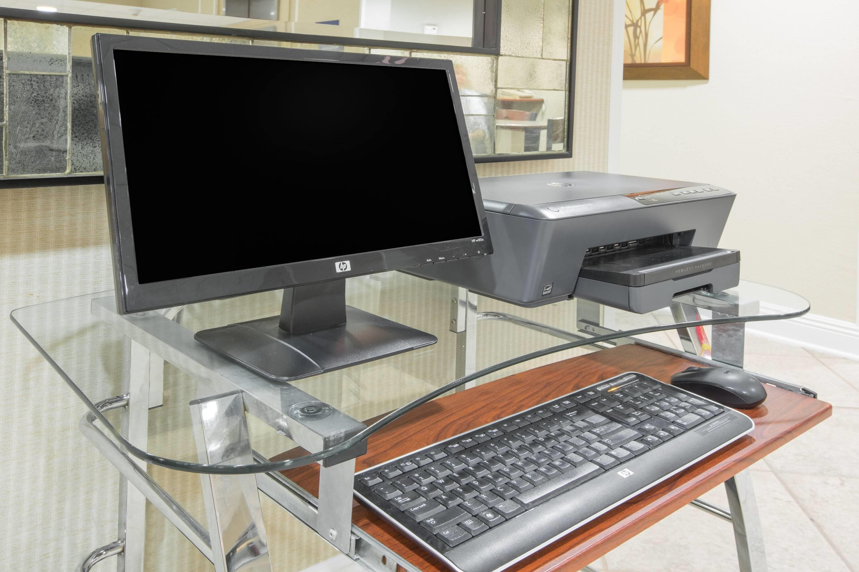A computer setup with a monitor, keyboard, and printer on a glass desk.