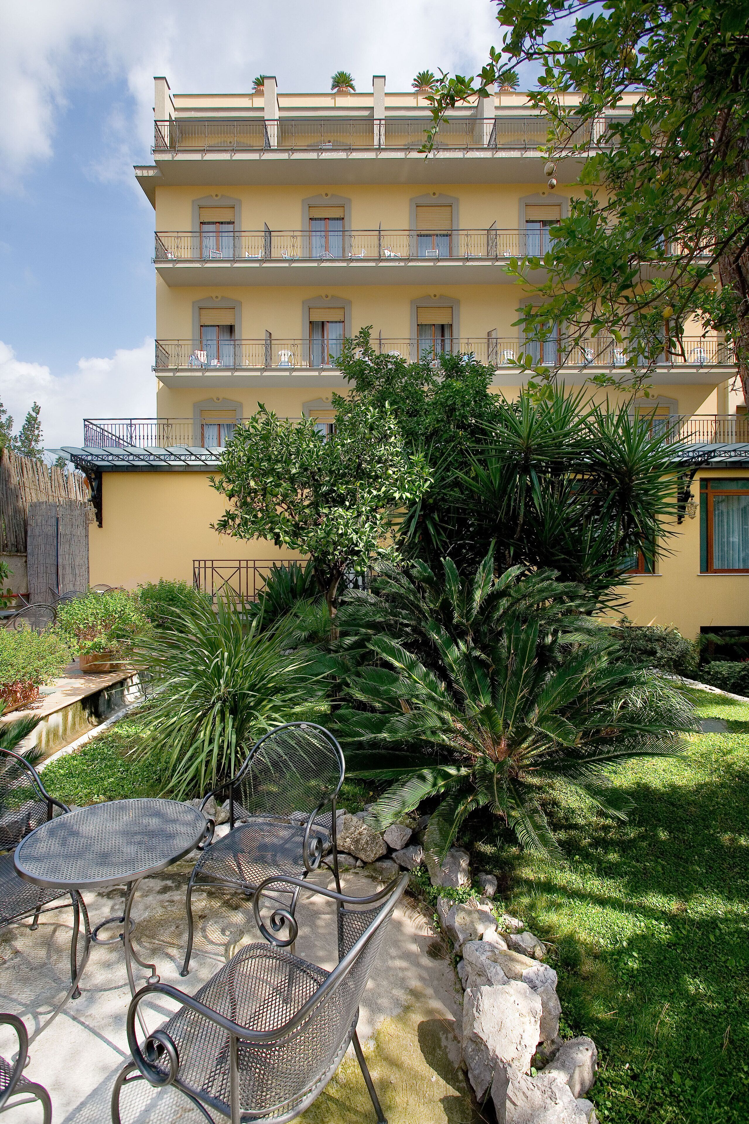 Un edificio amarillo con balcones, un jardín con plantas tropicales y mobiliario exterior.
