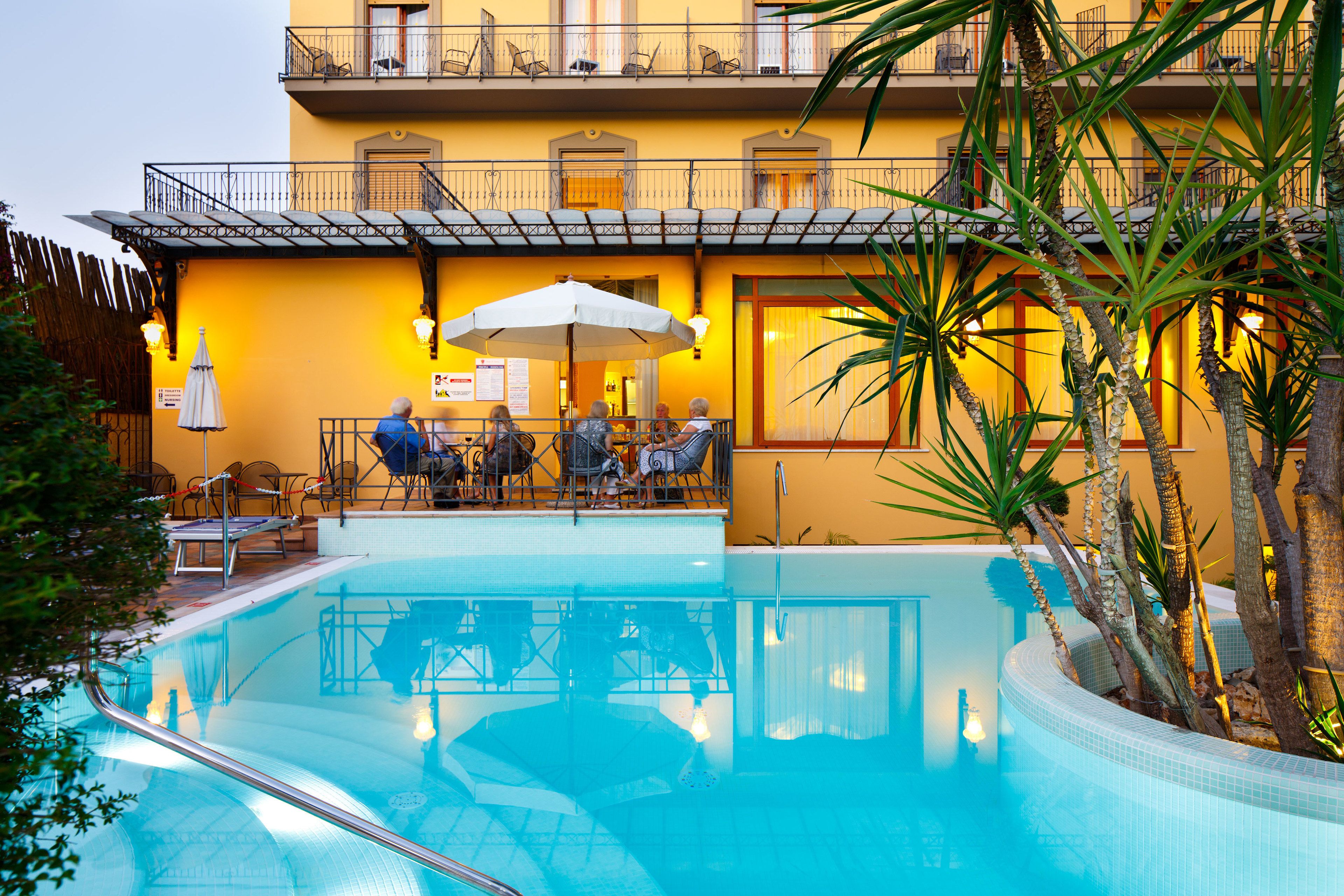 Ein Poolbereich mit Esstisch und Stühlen, einem weißen Sonnenschirm und einem Gebäude mit Balkonen.