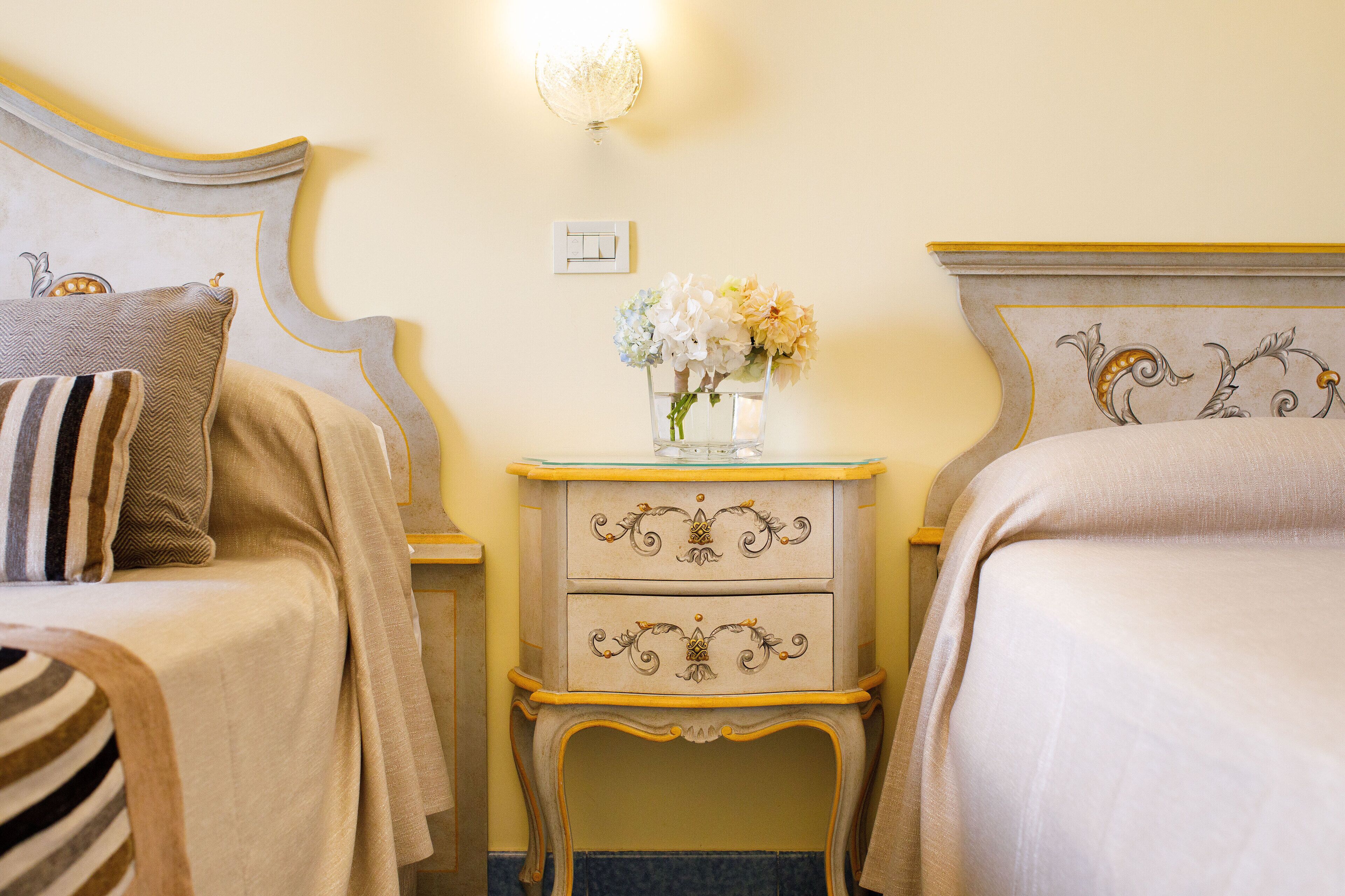A hotel room with two beds, a nightstand with a vase of flowers, and a wall-mounted light fixture.
