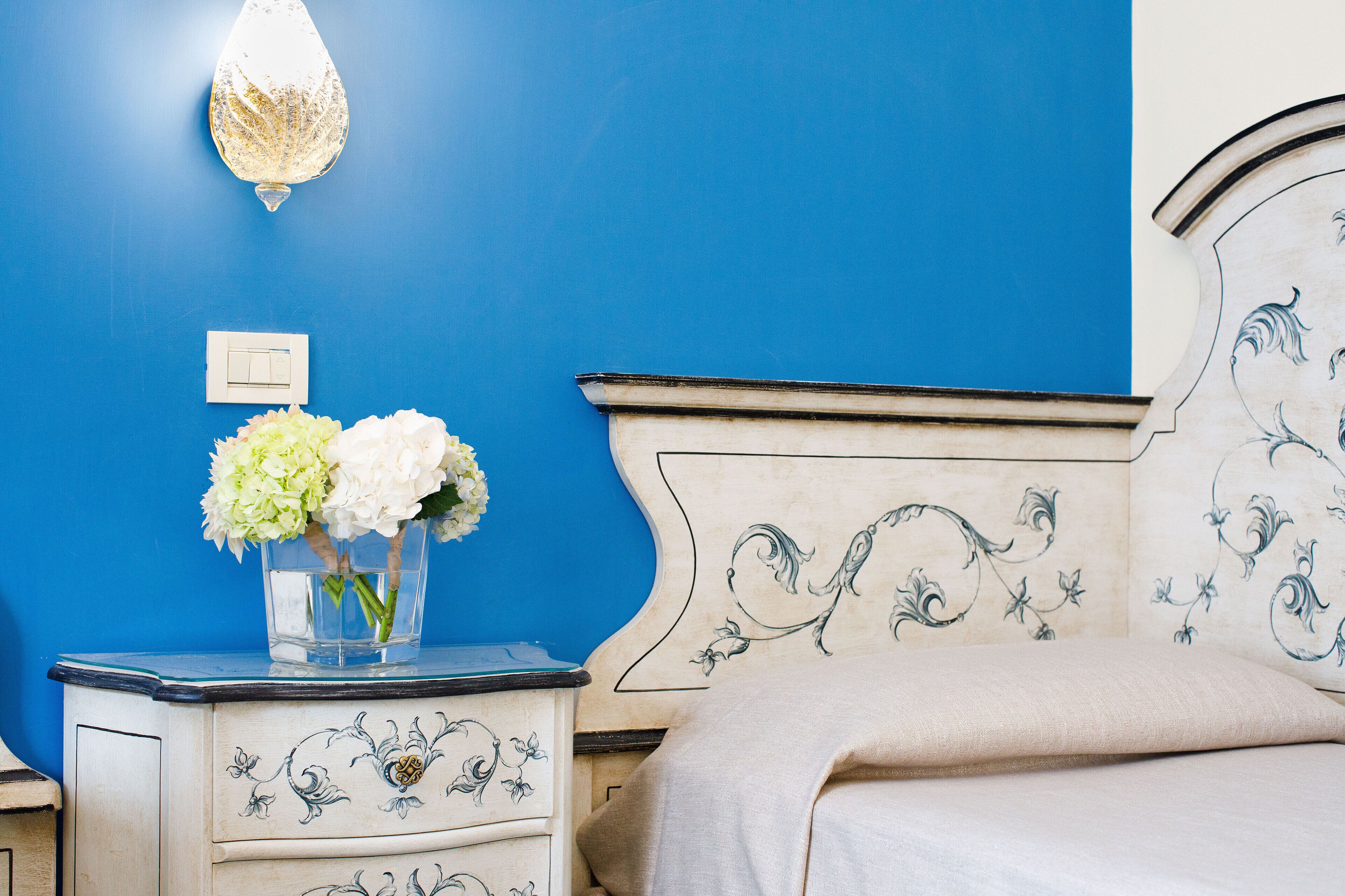 A bedroom with a blue wall, a white bedside table with a vase of flowers, and a bed with a white headboard.
