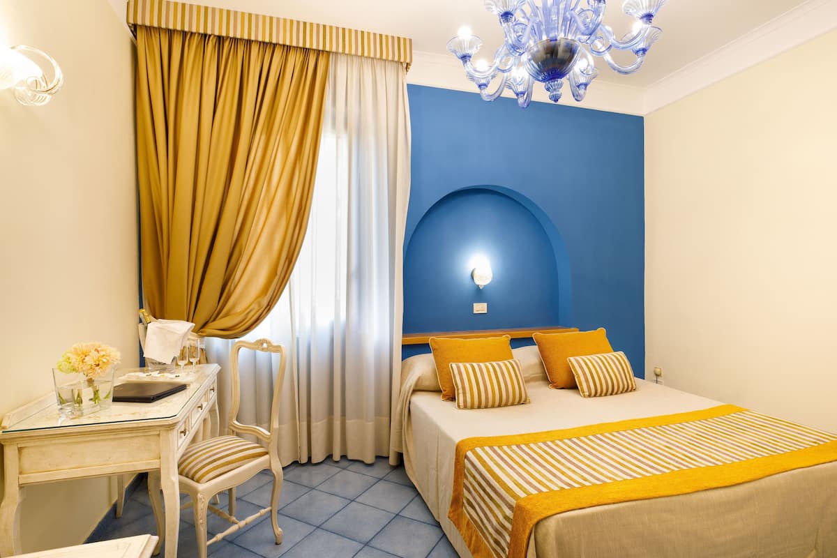 Habitación de hotel con cama, escritorio, sillón, araña y pared decorada en azul.
