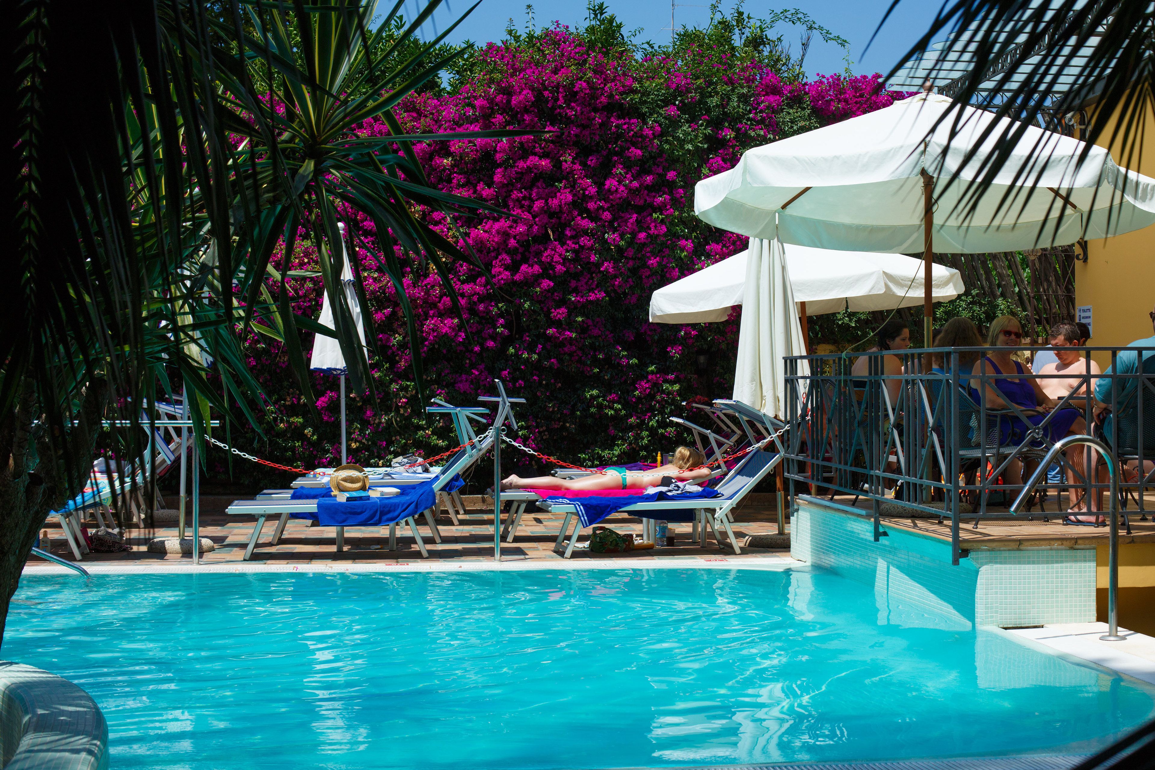 Área de piscina com espreguiçadeiras e guarda-sóis, cercada por flores rosa vibrantes e vegetação.