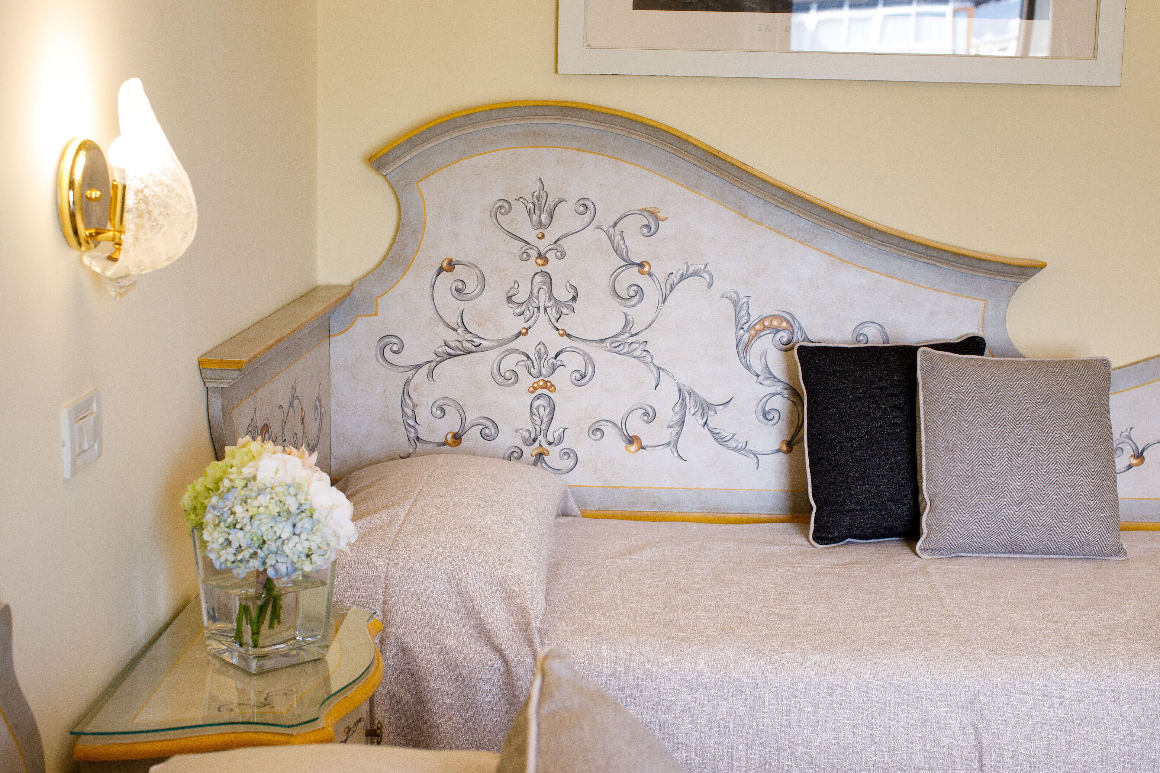 Una cama bien hecha con un cabecero decorativo, una mesita de noche con superficie de vidrio y un florero con flores.