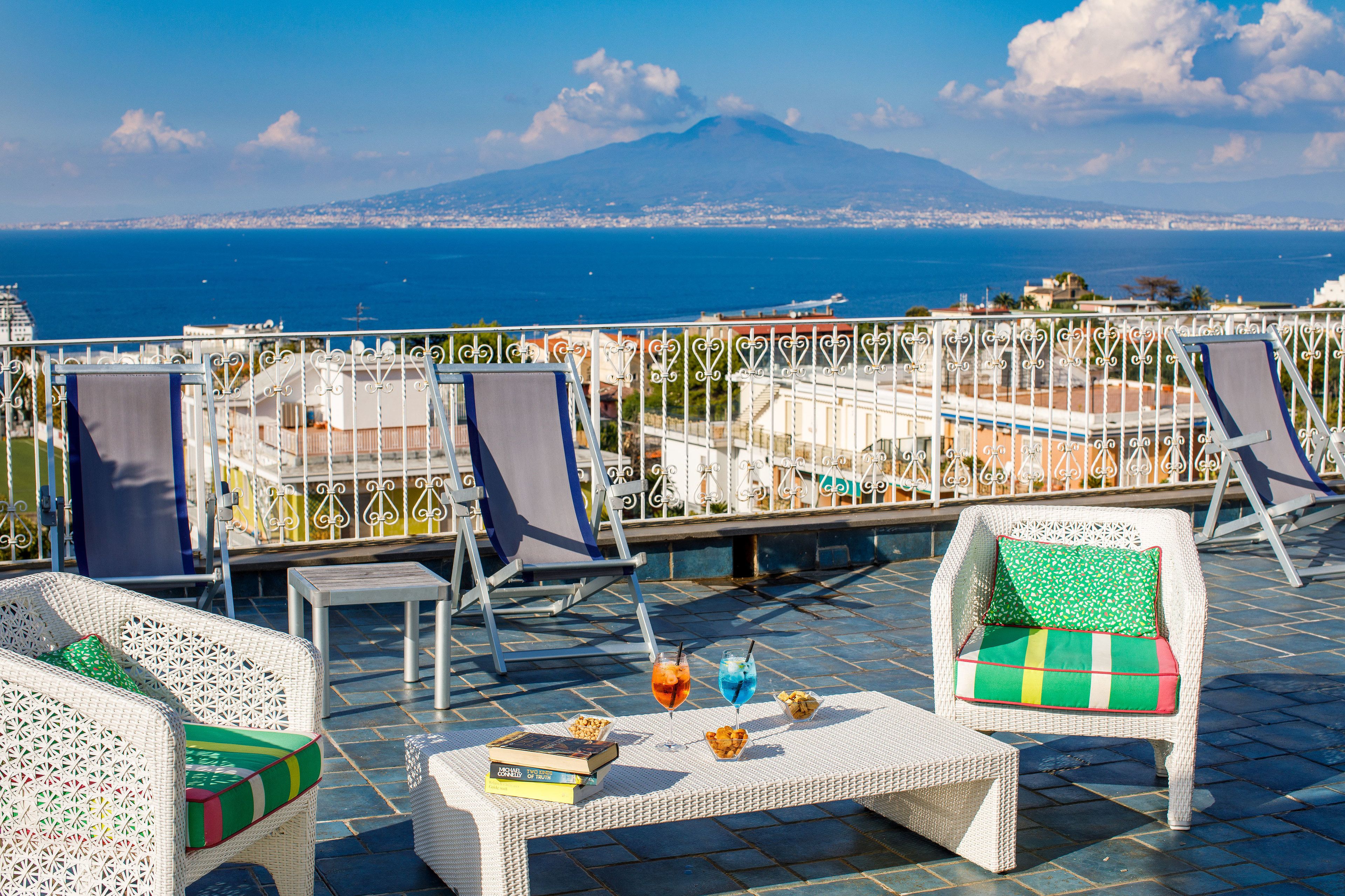 A terrace with wicker furniture, a table with books and snacks, and a view of the sea and a mountain.