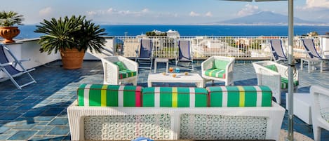 Una terraza con mobiliario de mimbre blanco, cojines a rayas verdes y blancas, y vista al mar.