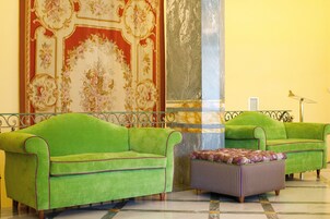 A room with two green sofas and a purple ottoman, a large floral tapestry, and a marble column.
