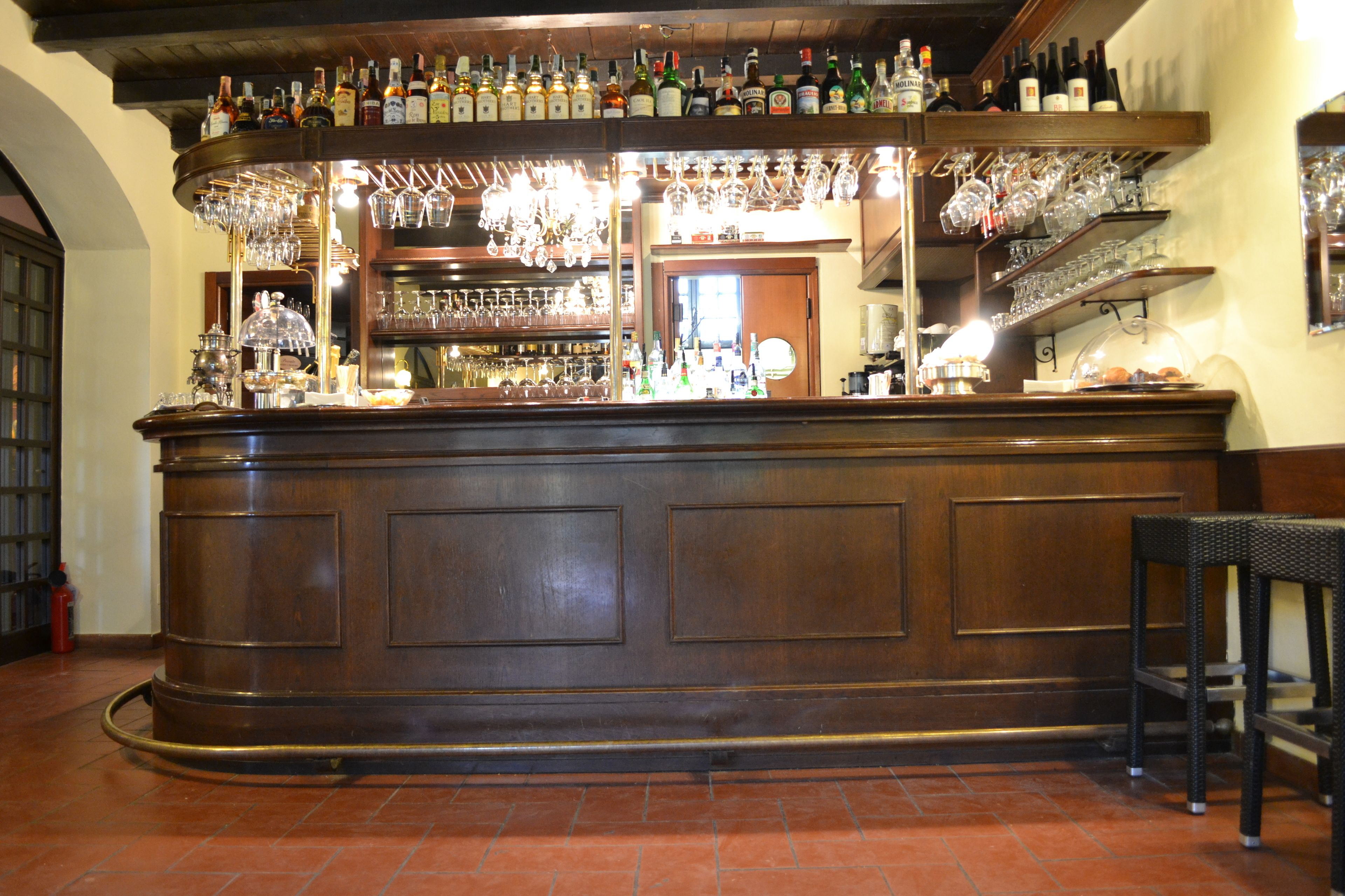 Zona de bar con mostrador de madera, estantes con botellas y vasos, y suelo de baldosas.