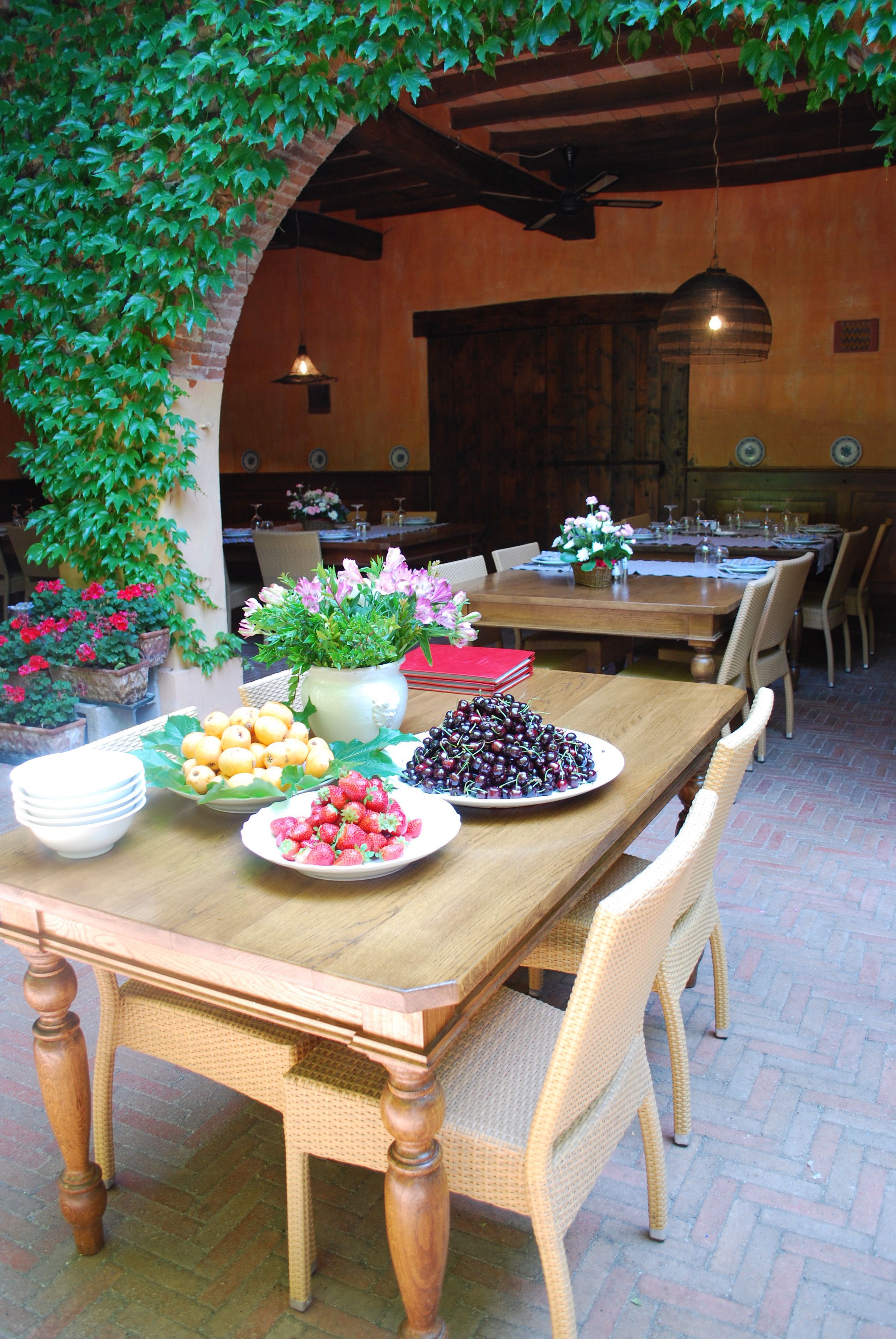 Una mesa de comedor de madera con sillas de mimbre, rodeada de plantas en macetas y una variedad de frutas y flores.