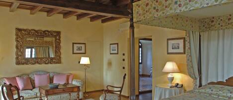 A room with a four-poster bed, a sofa, a chair, a table, and a mirror. There are two lamps on the table and a picture frame on the wall.