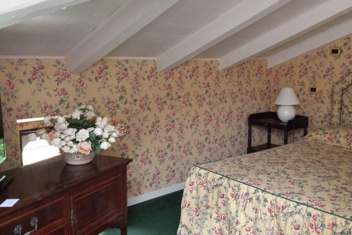 A bedroom with floral wallpaper, a bed with a floral bedspread, a nightstand with a lamp, and a dresser with a vase of flowers.