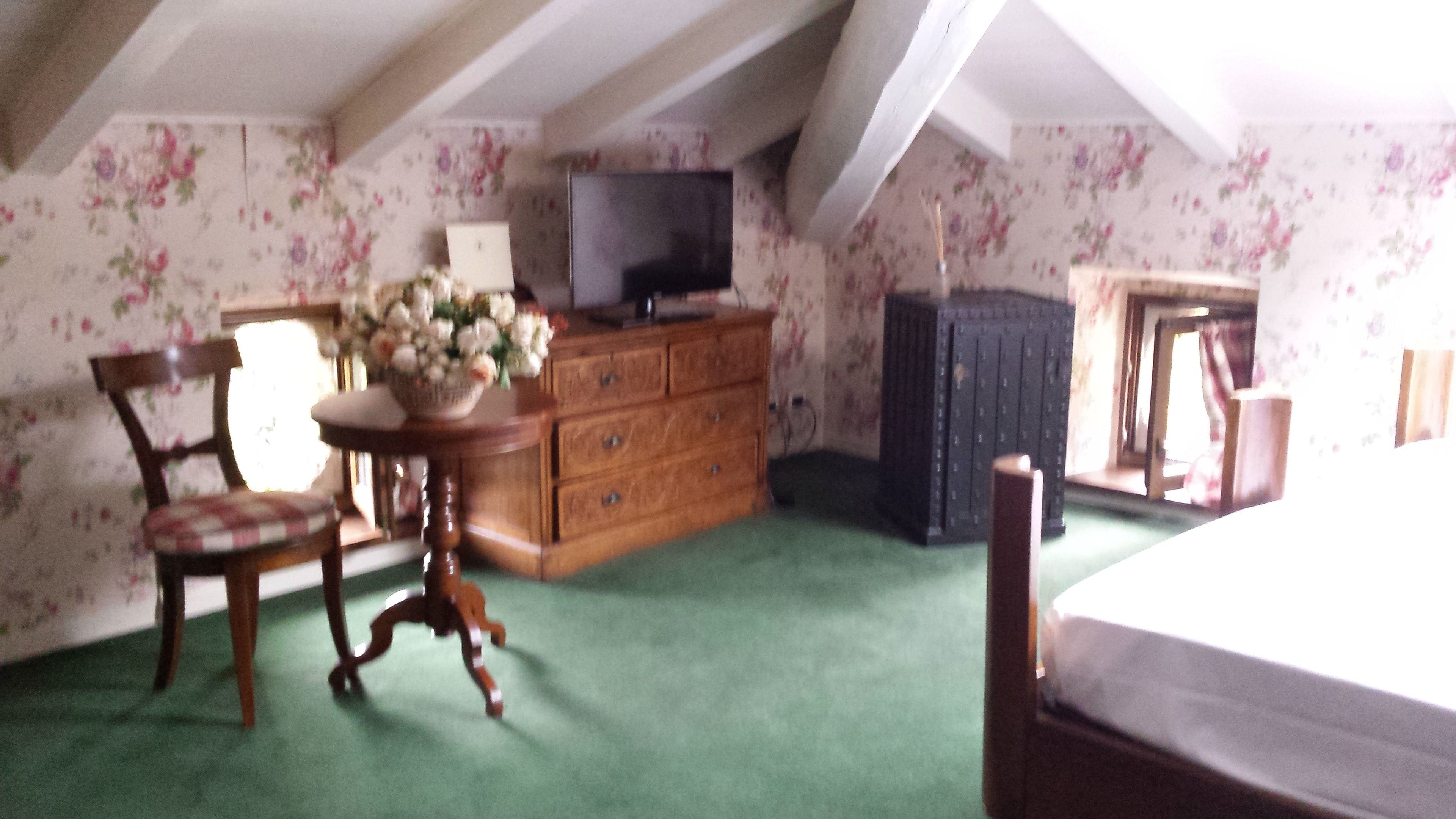A room with a wooden dresser, a television on top, a chair with a checkered cushion, and a floral wallpaper.