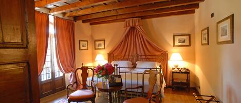 Ein Schlafzimmer mit Baldachinbett, zwei Stühlen, einem kleinen Tisch mit Blumenvase und einem Nachttisch mit Lampe.