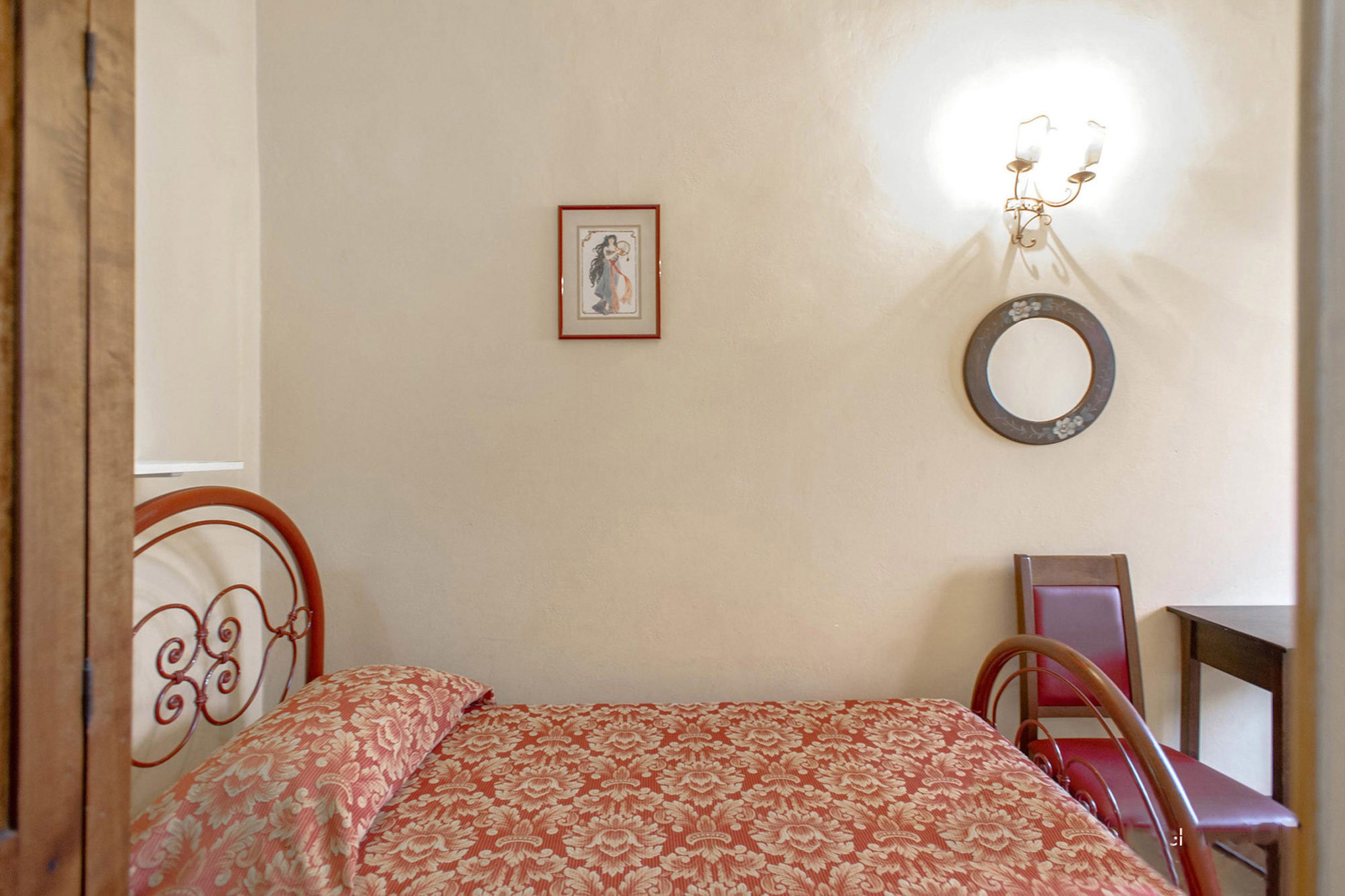 A bedroom with a bed, a chair, a small table, a wall-mounted light fixture, and a framed picture.