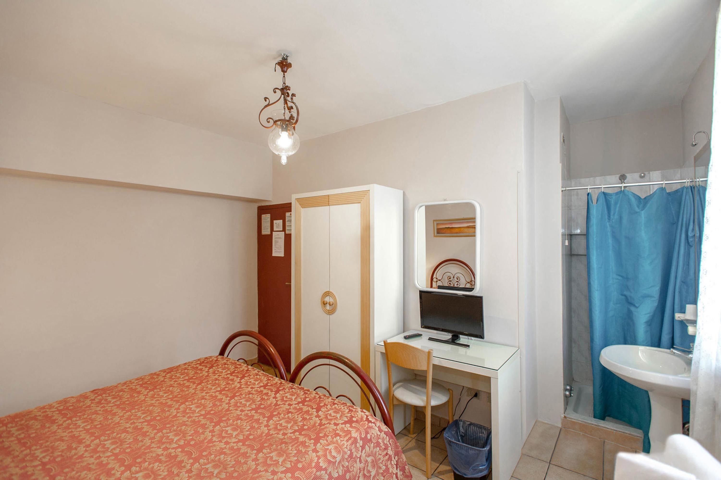 A bedroom with a bed, a desk with a computer, a mirror, and a bathroom with a sink and shower curtain.