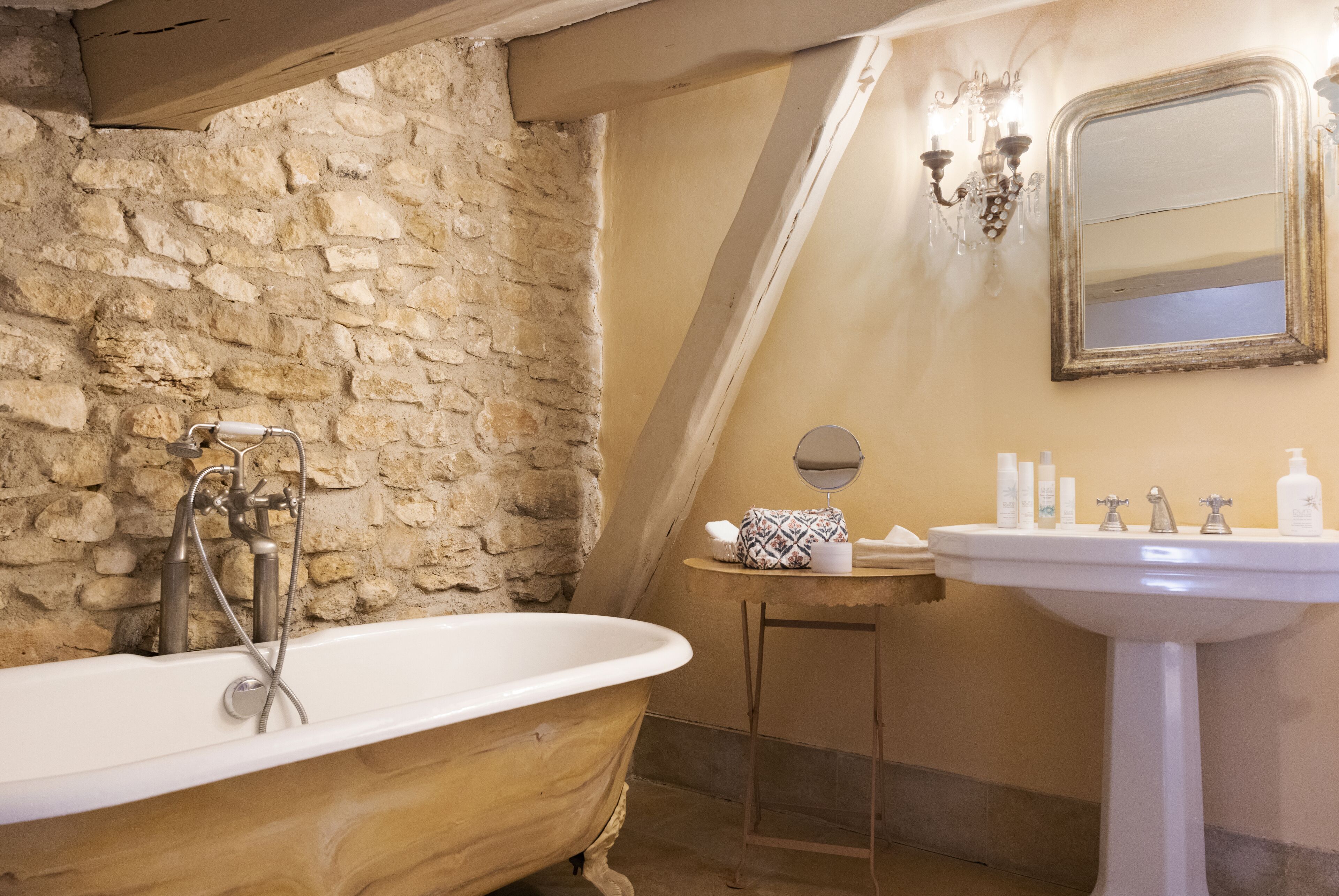 A bathroom with a clawfoot bathtub, a stone wall, a small vanity with a mirror, and toiletries on the sink.