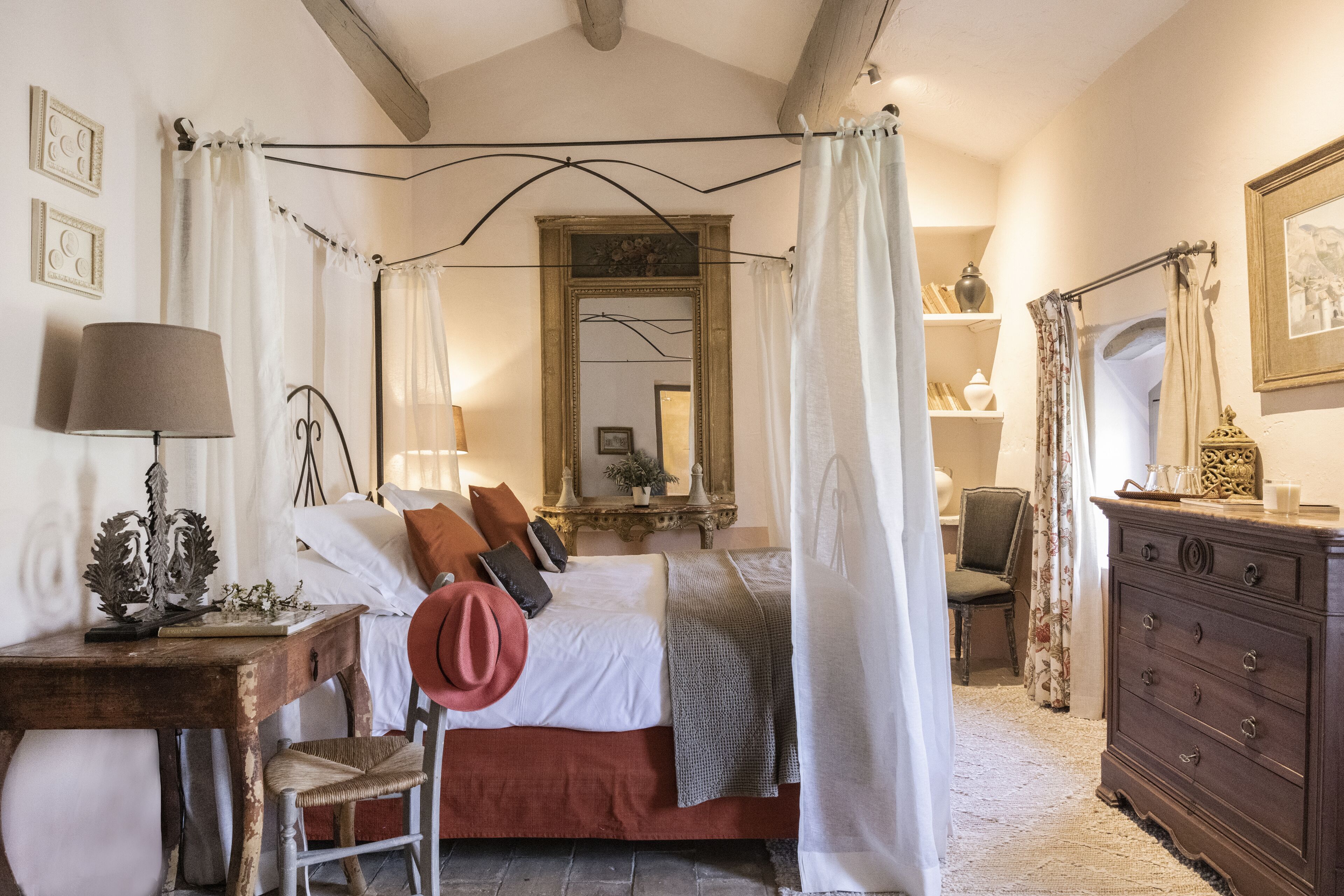 A bedroom with a four-poster bed, a wooden dresser, a chair, and a desk with a lamp.