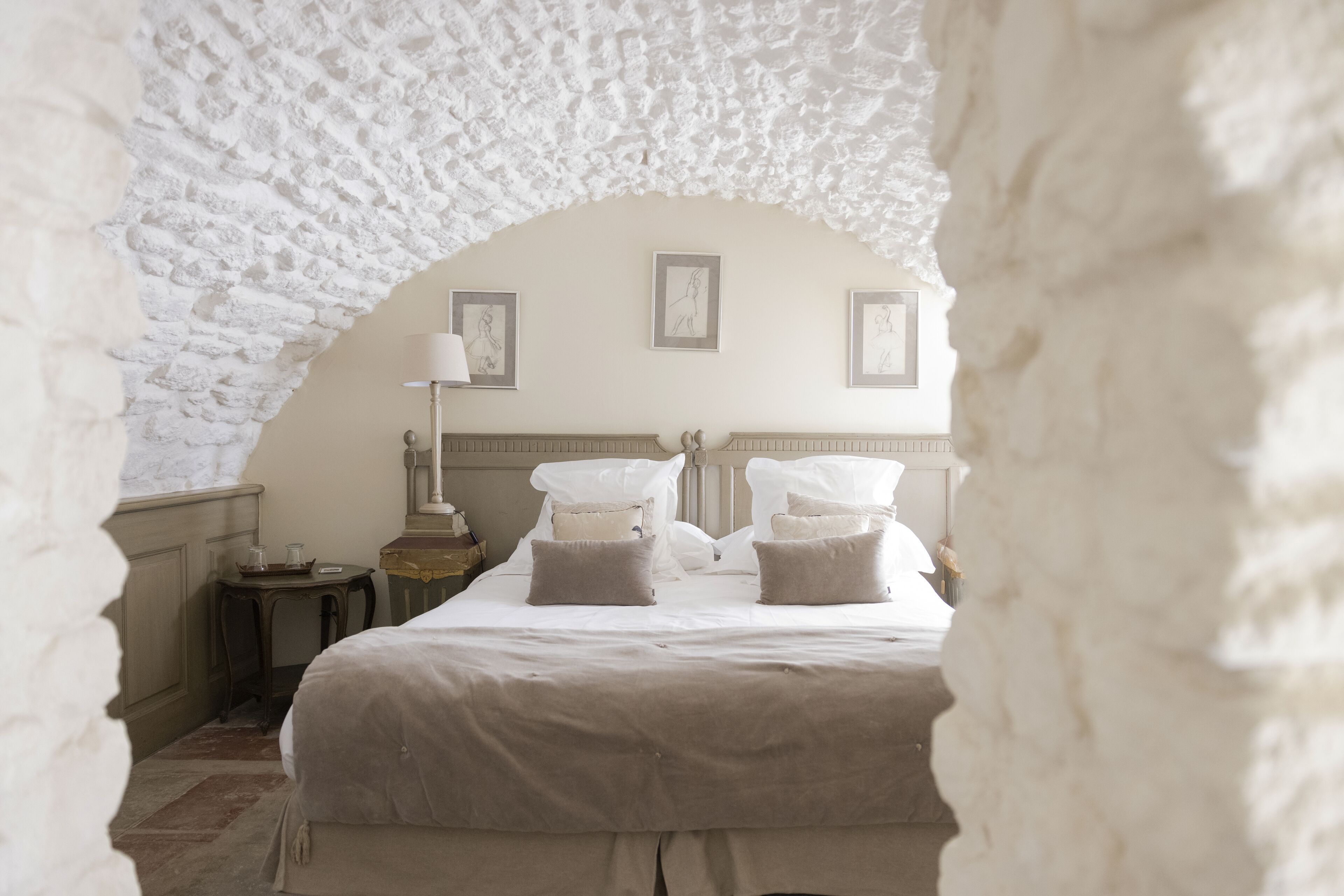 Une pièce dotée d’une arche en pierre, d’un lit avec du linge de maison blanc, d’une table de chevet et de tableaux encadrés accrochés au mur.