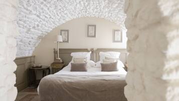 Une pièce dotée d’une arche en pierre, d’un lit avec du linge de maison blanc, d’une table de chevet et de tableaux encadrés accrochés au mur.