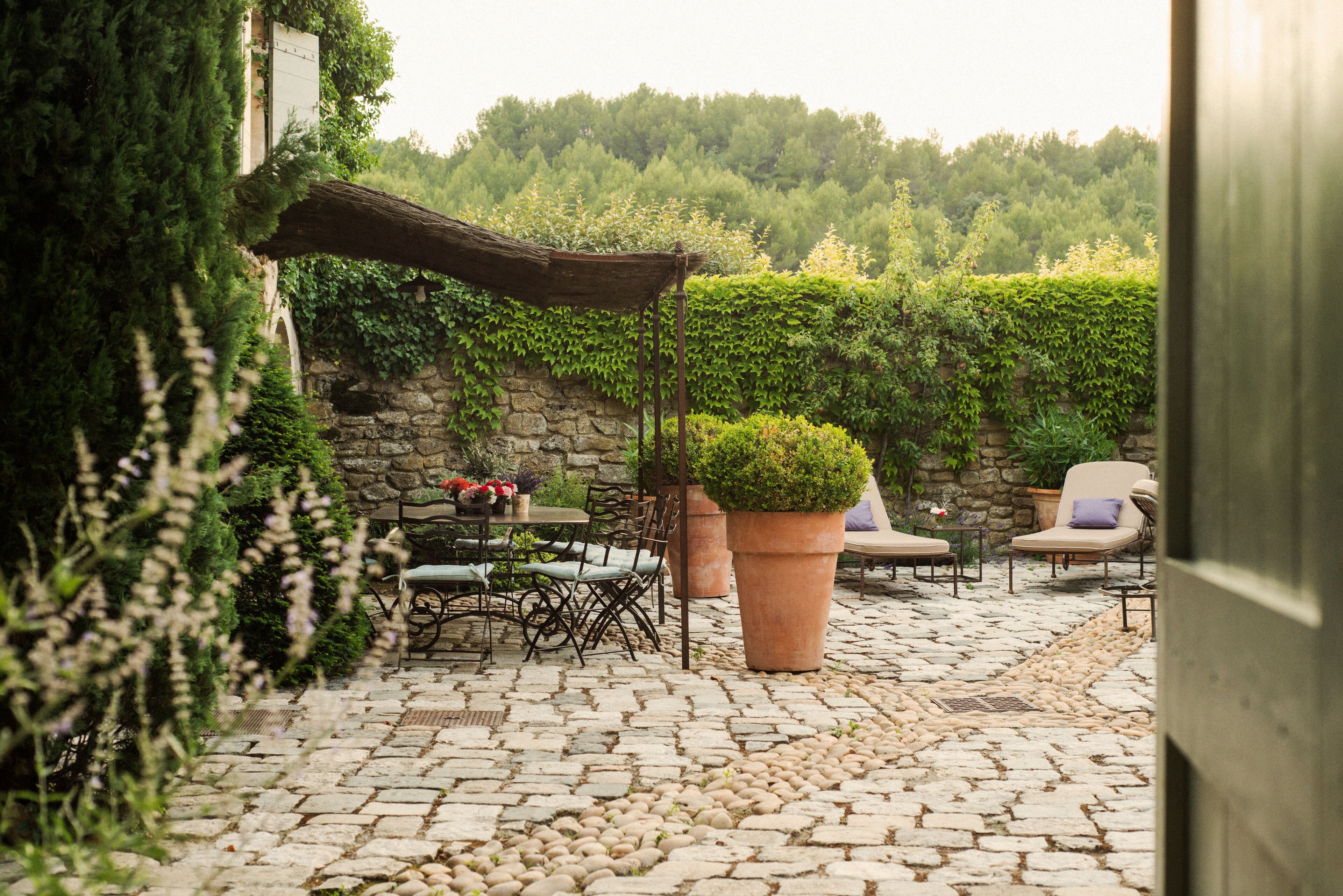An outdoor patio with stone flooring, a table with chairs, a large potted plant, and a covered seating area.