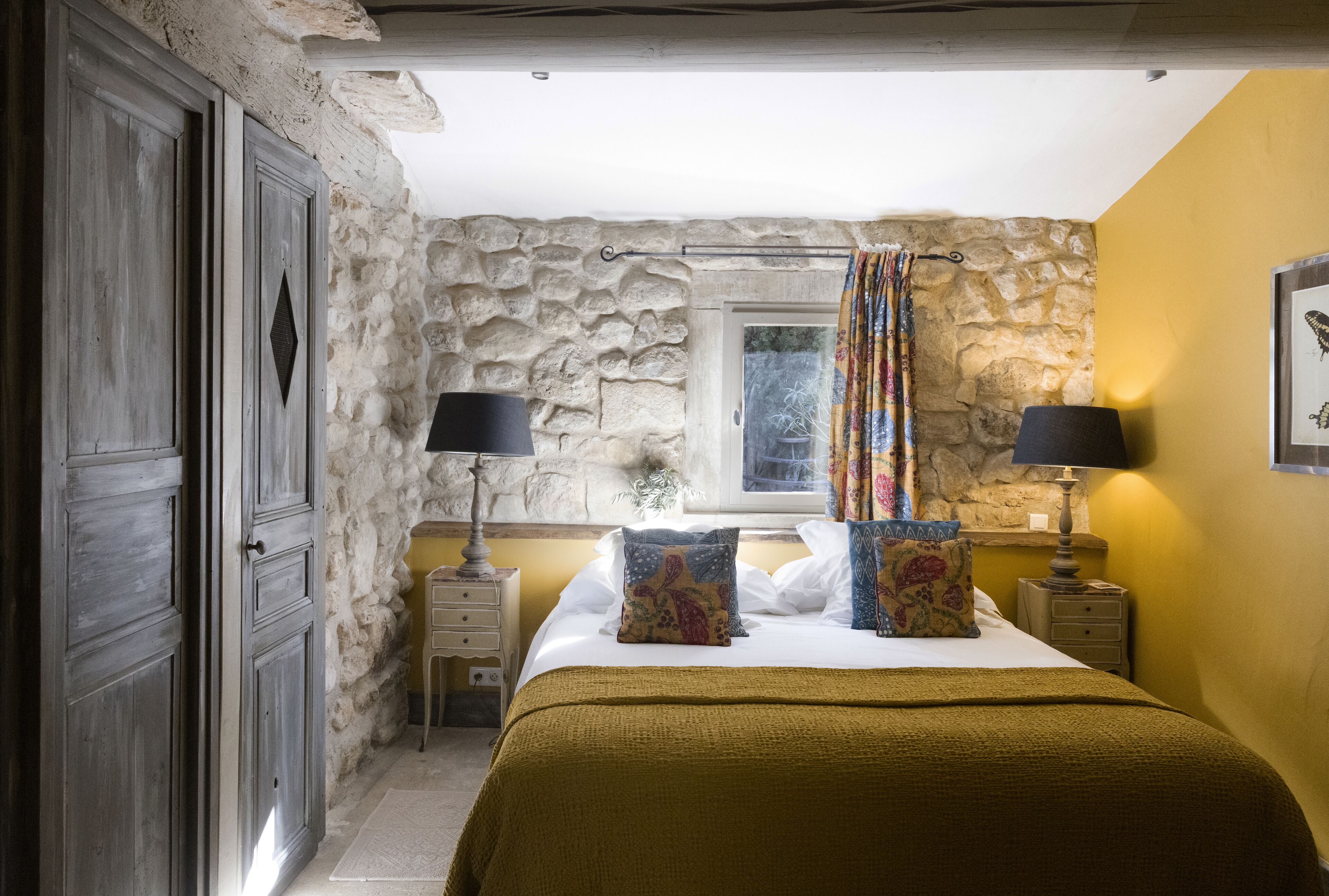 Ein Schlafzimmer mit Steinkamin, einem Bett mit vielen Kissen, zwei Nachttischen mit Lampen und einem Fenster mit Vorhängen.