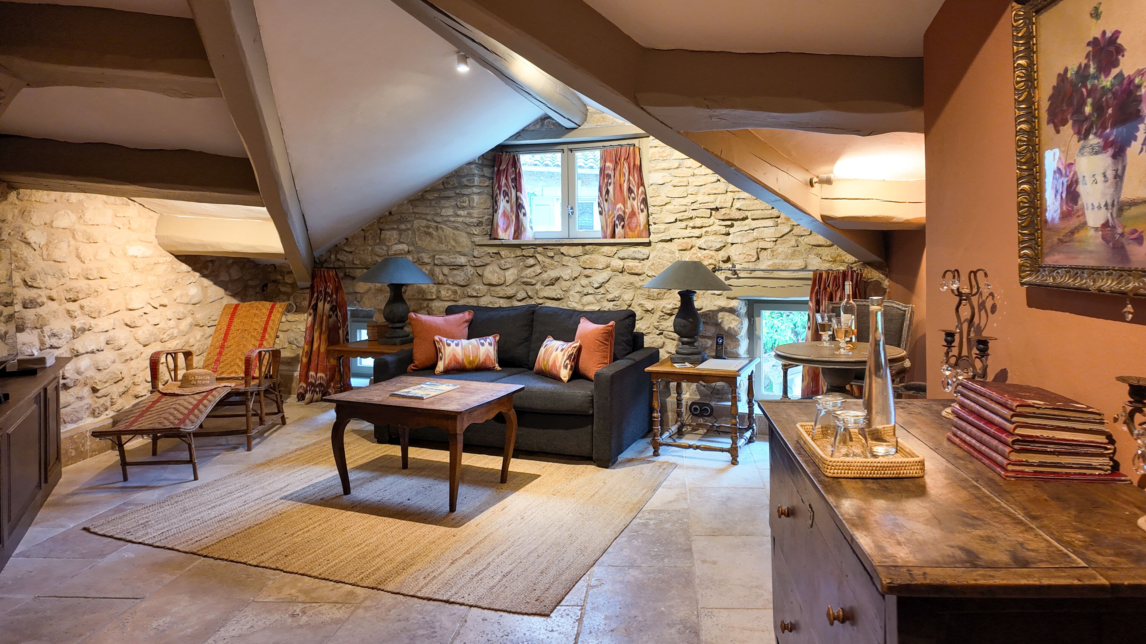 A cozy loft-style room with a stone wall, a dark sofa with patterned cushions, a wooden coffee table, and a dining table with books and a candle.