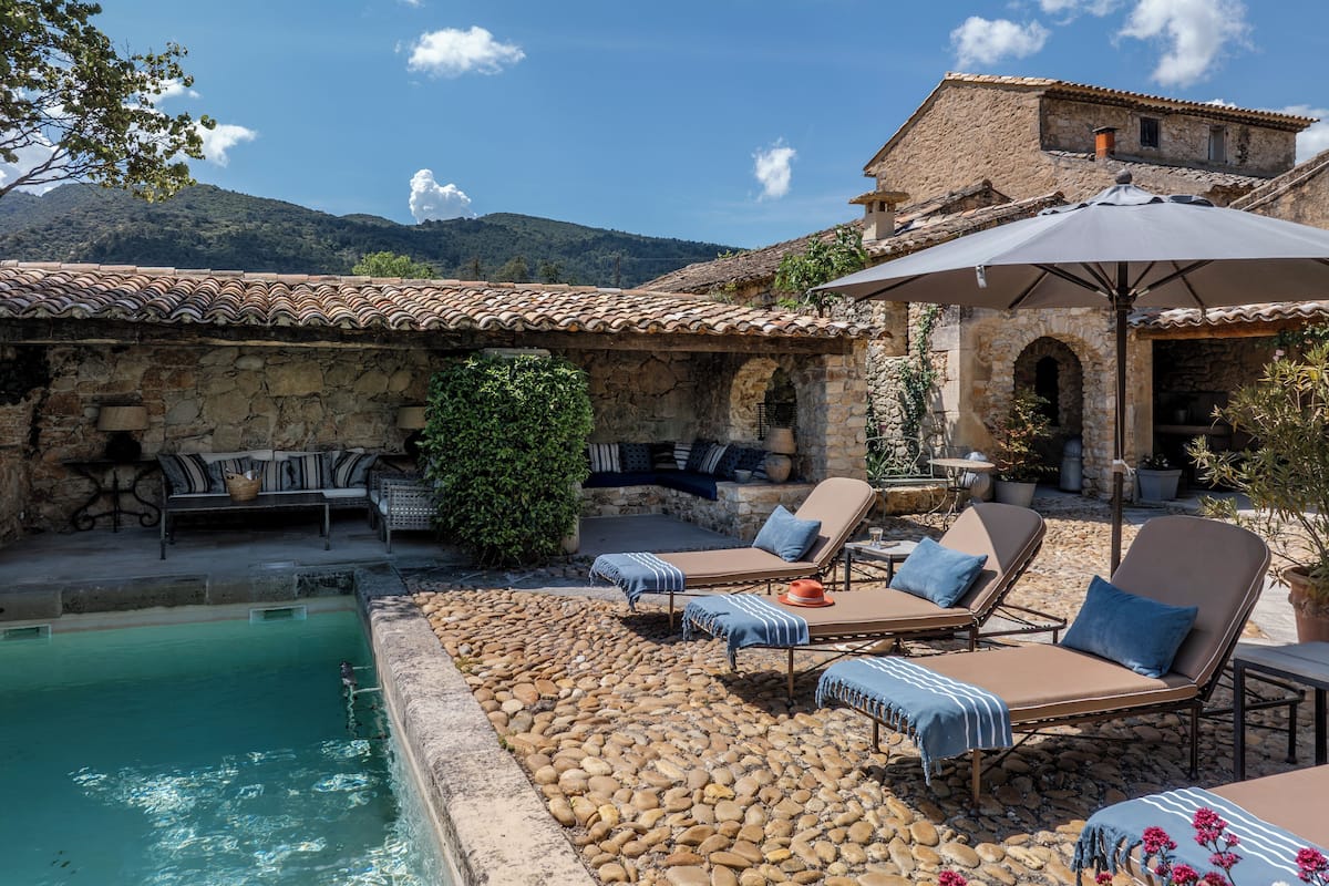Un espace aménagé au bord de la piscine, doté d’un sol en pierre, de chaises longues et d’un bâtiment en pierre surmonté d’un toit en tuiles.