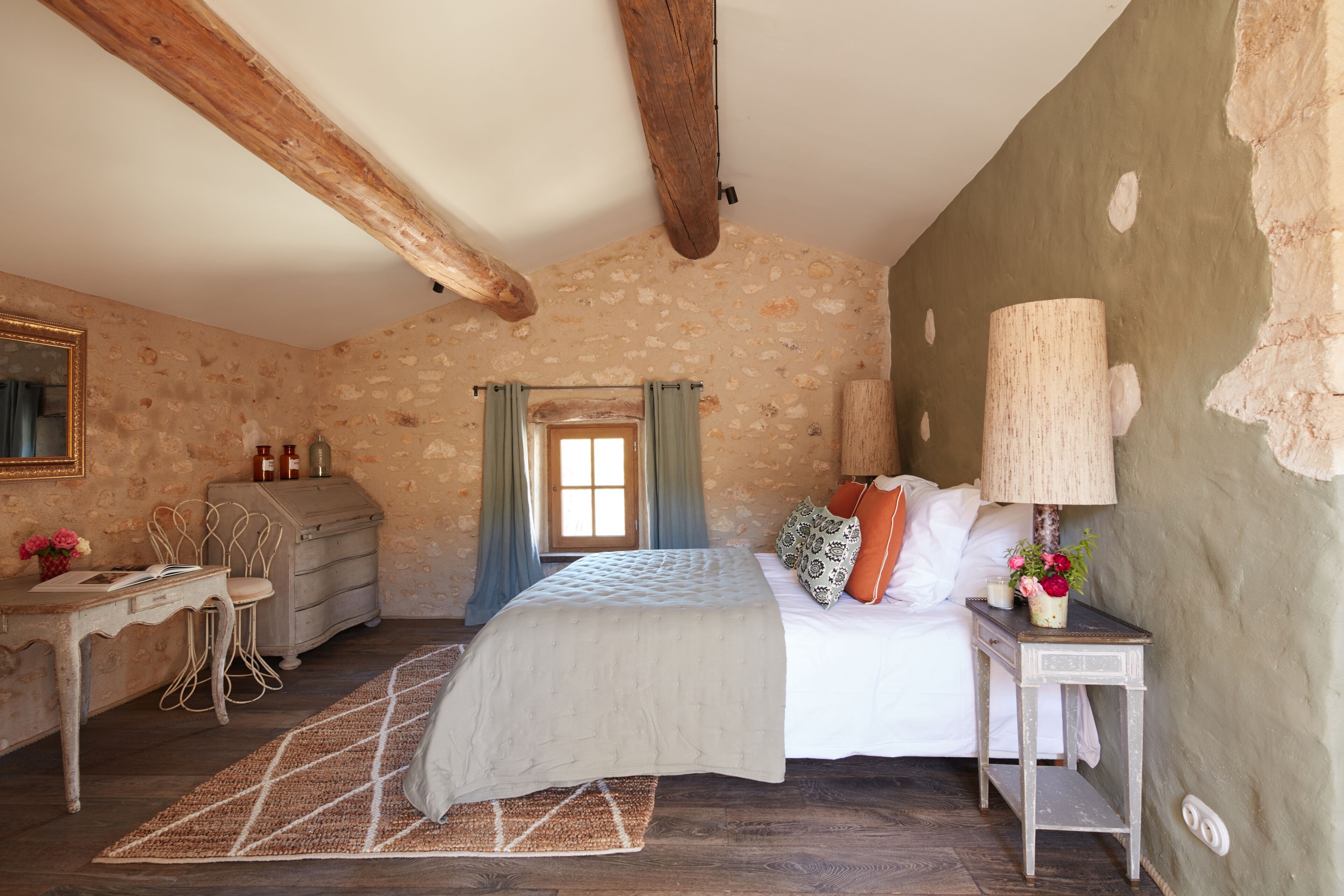 A rustic bedroom with exposed wooden beams, a bed with white linens, a desk with a chair, and a small table with a lamp and flowers.