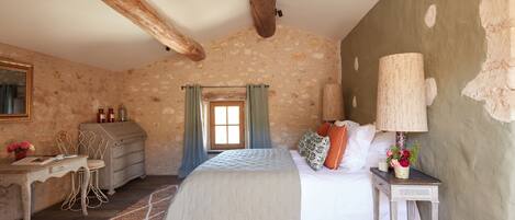 A rustic bedroom with exposed wooden beams, a bed with white linens, a desk with a chair, and a small table with a lamp and flowers.