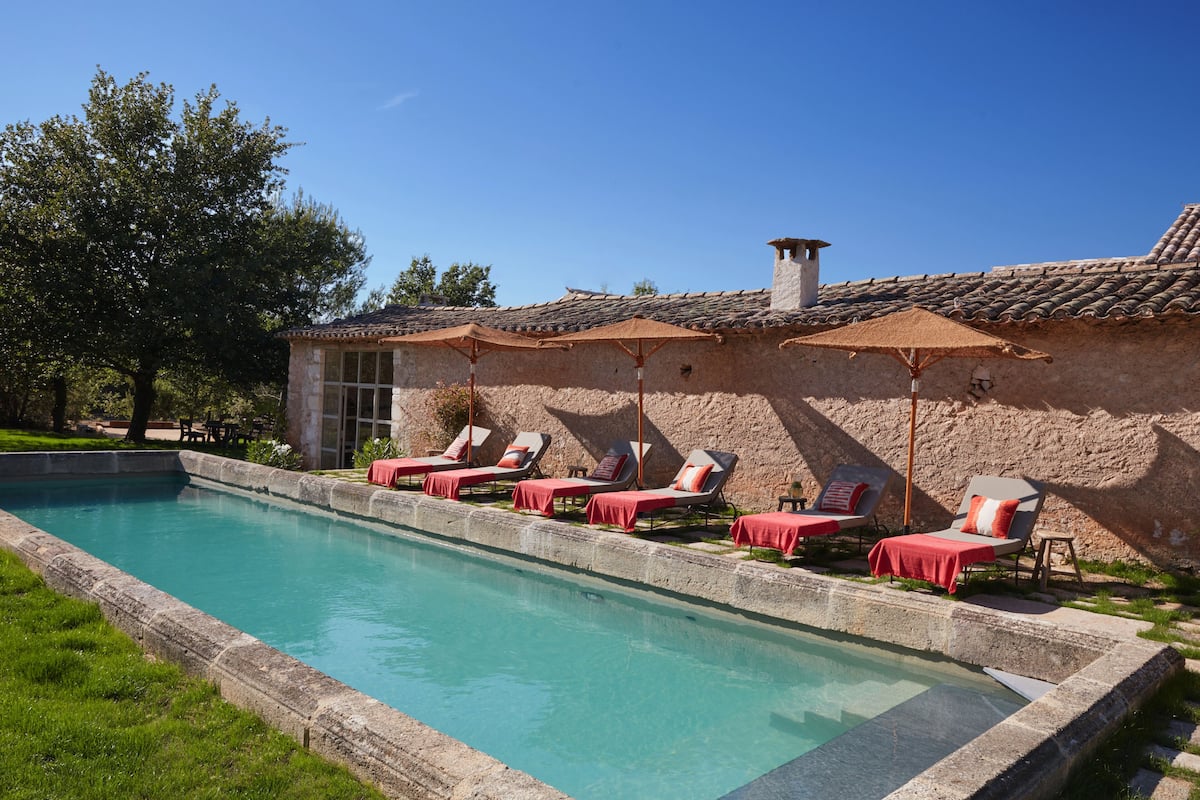 Une piscine entourée d’un mur de pierre et d’un arbre, avec des chaises longues et des parasols à proximité.