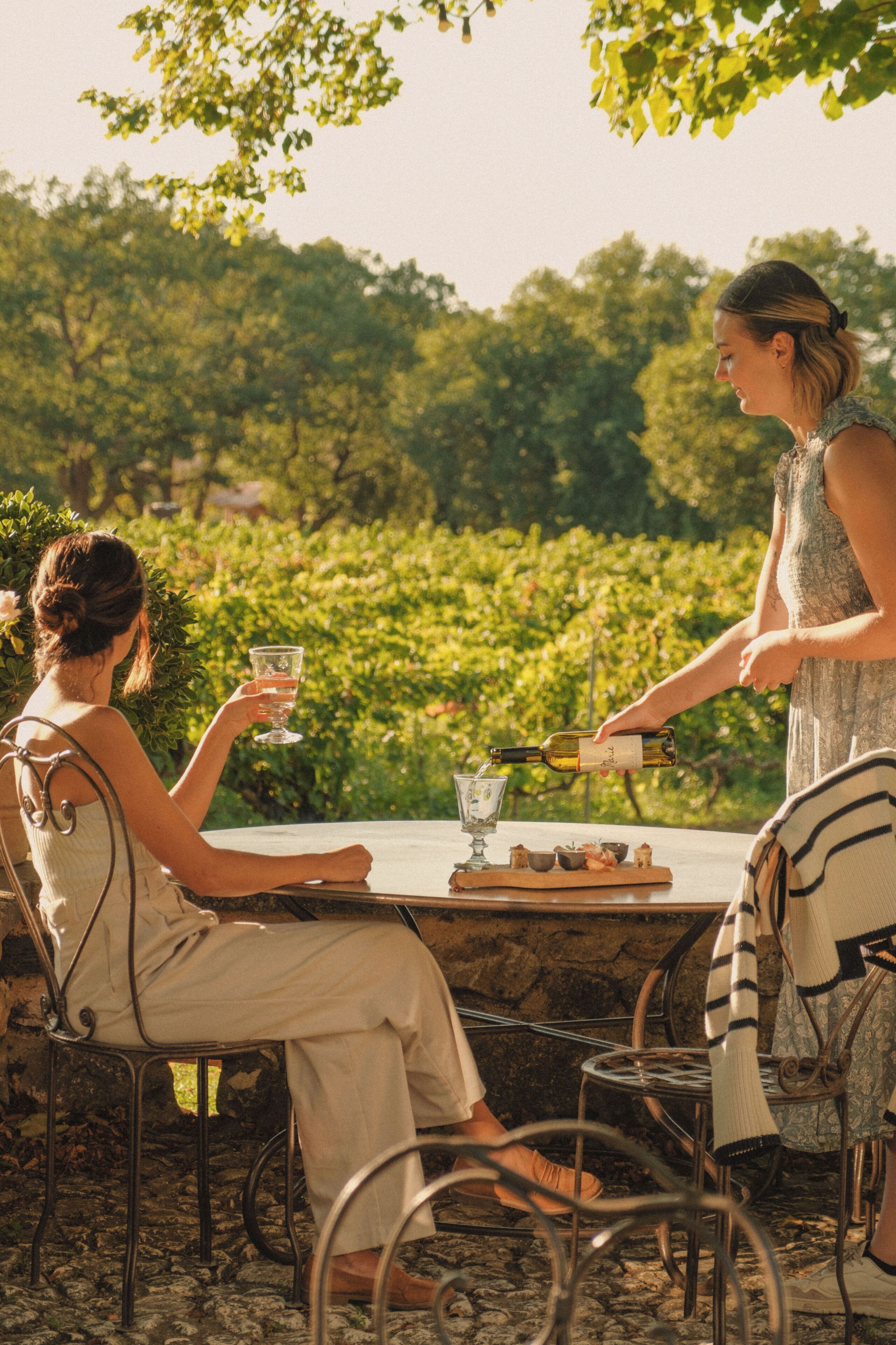 Two people seated at an outdoor table with a wine bottle and glasses, surrounded by greenery.