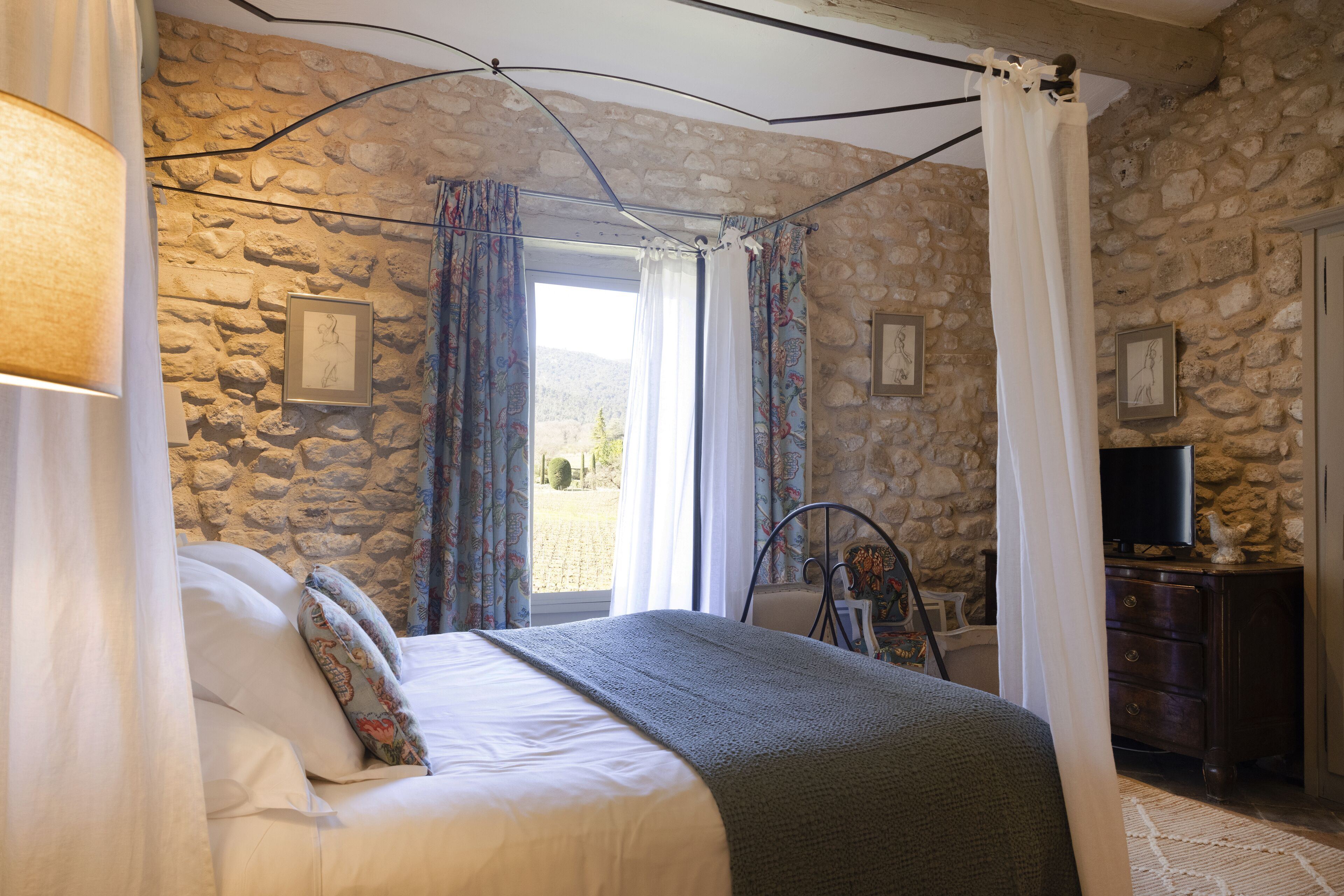 A bedroom with a stone wall, a bed with white linens, a television on a wooden cabinet, and a window with curtains.