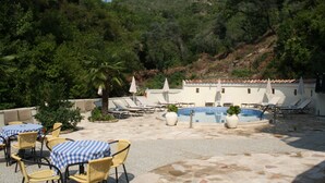 Zona de piscina con sillones reclinables y mesas, rodeada de vegetación y una colina al fondo.