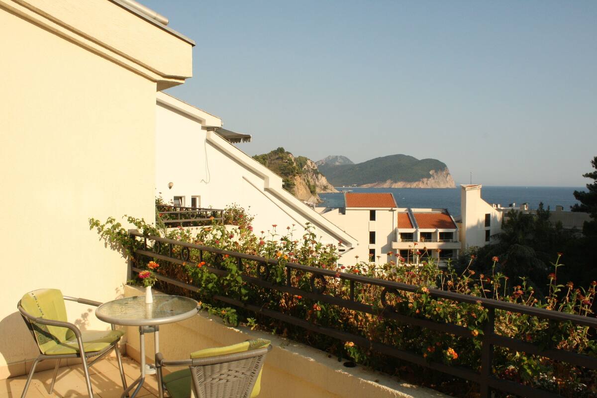 A balcony with a table and chairs, overlooking a coastal area with buildings and cliffs.