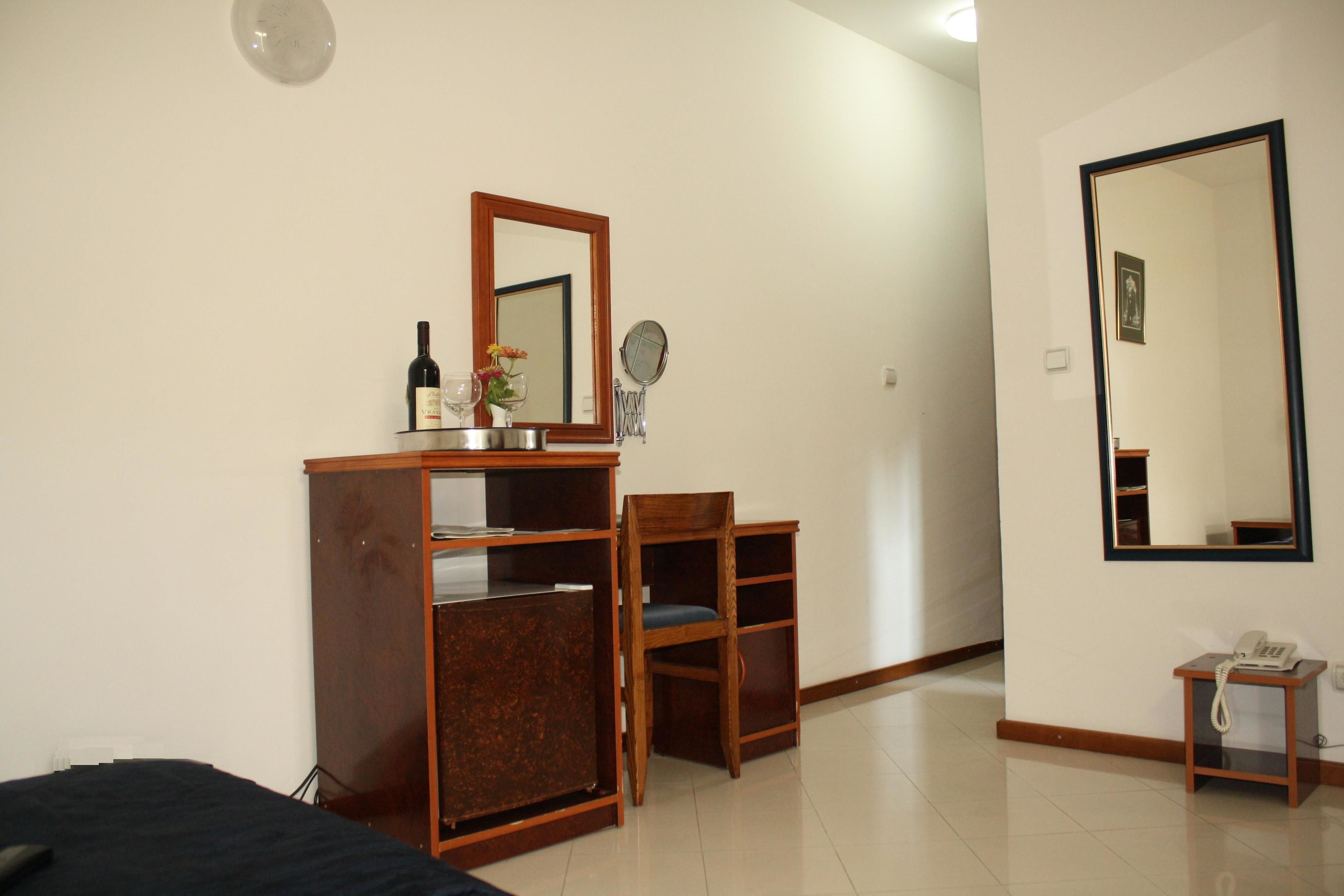 A bedroom with a bed, a wooden wardrobe, a desk with a mirror, a chair, and a bedside table with a phone.
