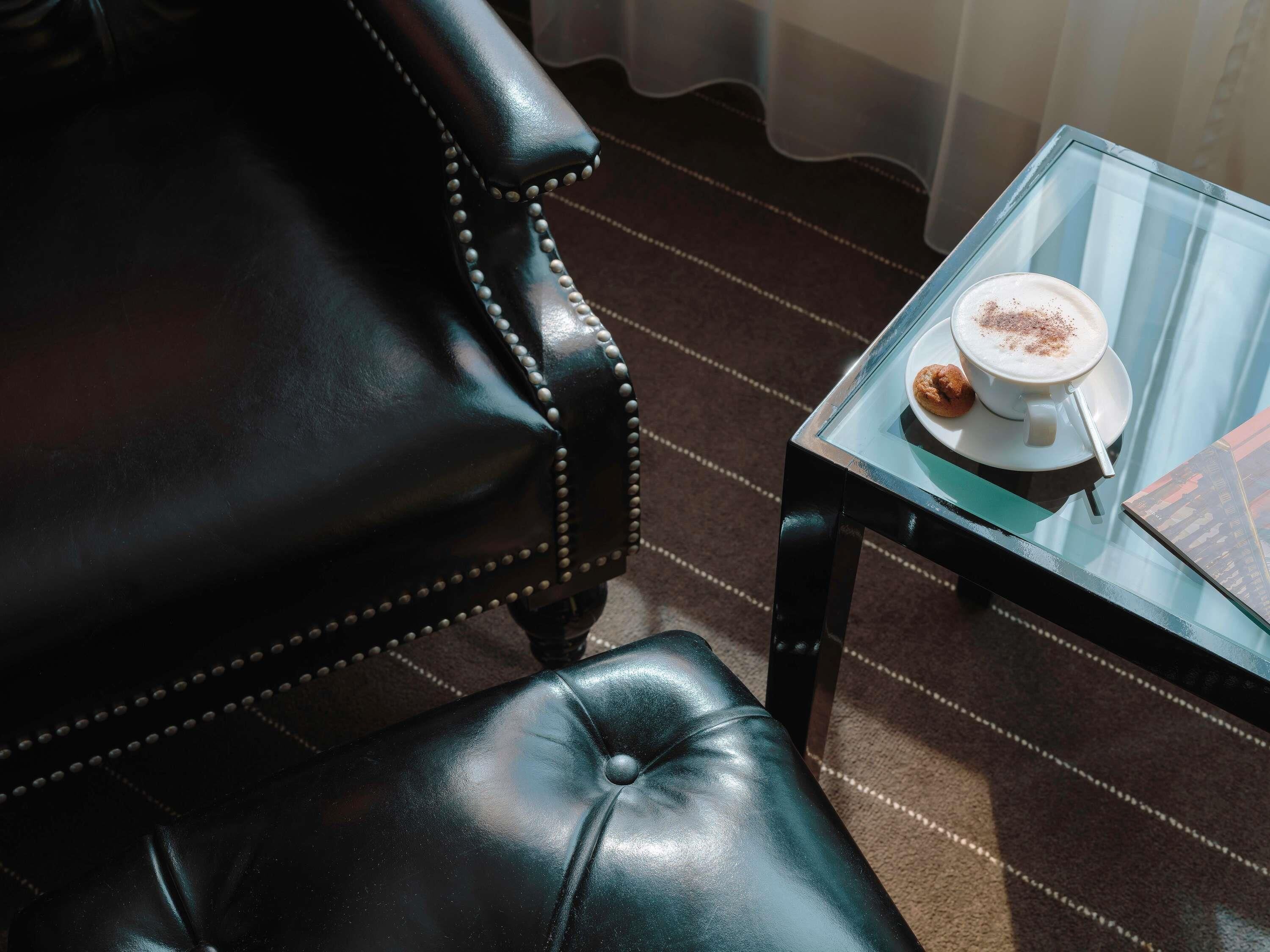 A black leather chair with a high backrest and a glass table with a cup of coffee and a pastry.