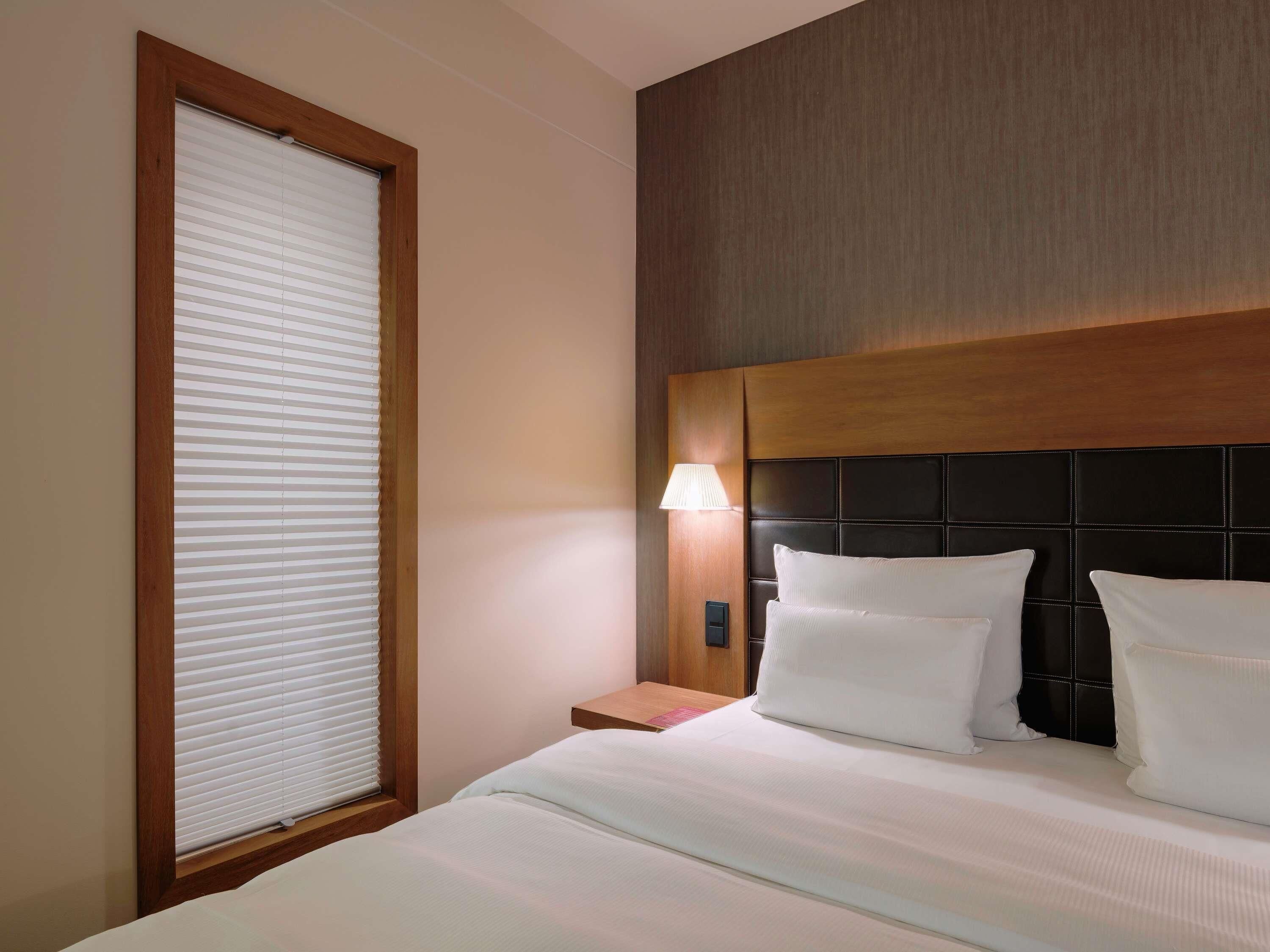 Een hotelkamer met een groot bed, een nachtkastje met een lamp en een raam met zonwering.