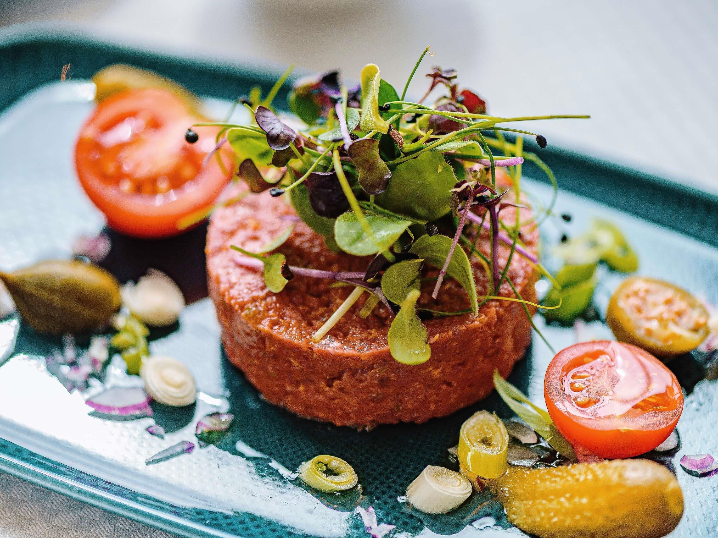 A dish with a central meat patty, garnished with greens and surrounded by sliced tomatoes and pickles.