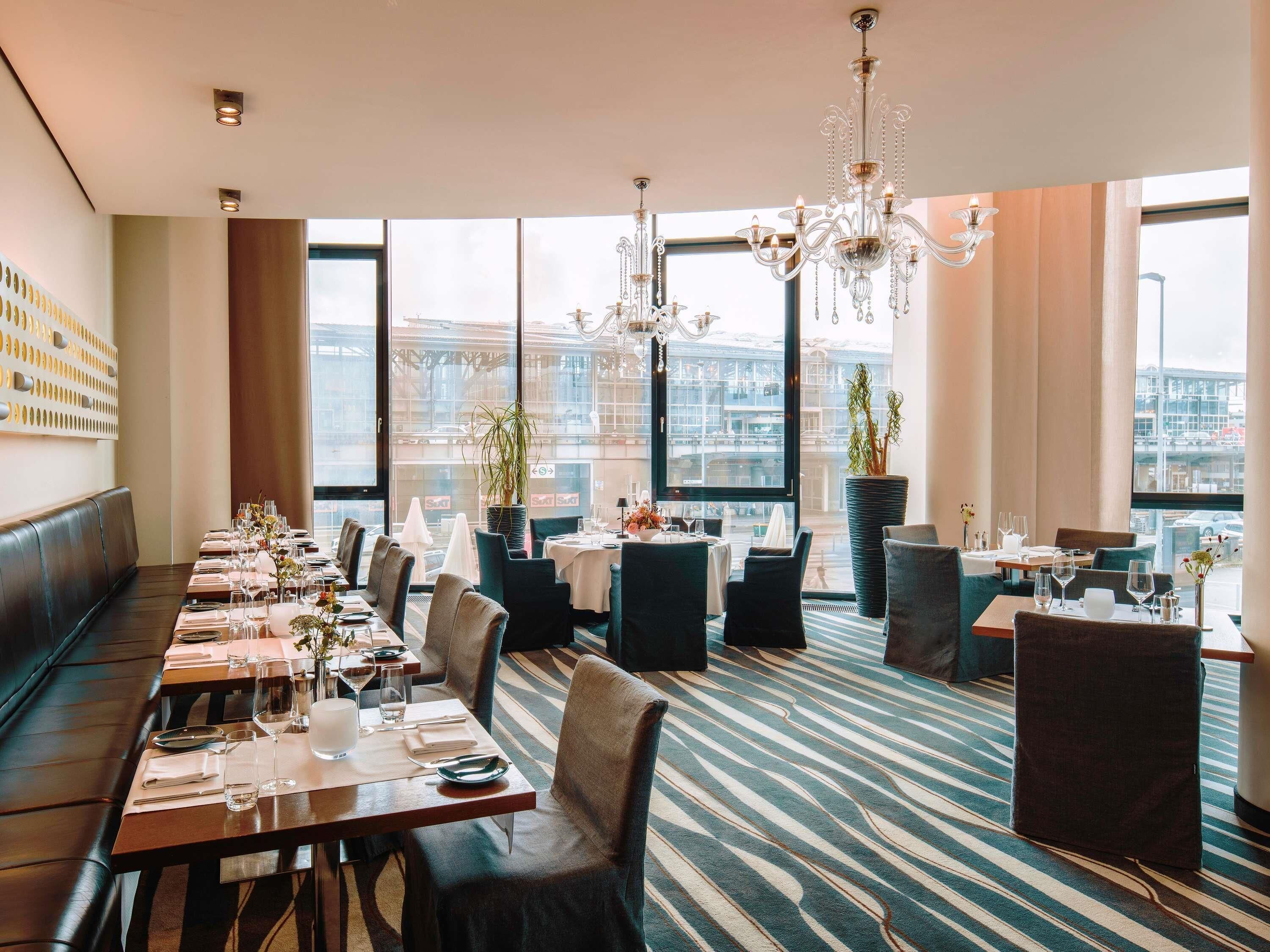 A restaurant with a long dining table, multiple chairs, a chandelier, and large windows offering a city view.