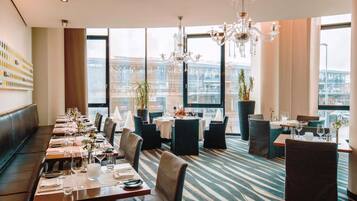 Un restaurant doté d’une longue table à manger, de nombreuses chaises, d’un lustre et de grandes fenêtres offrant une vue sur la ville.