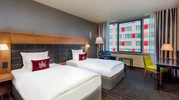 A hotel room with two beds, a desk, a chair, and a view of a building with red and white windows.