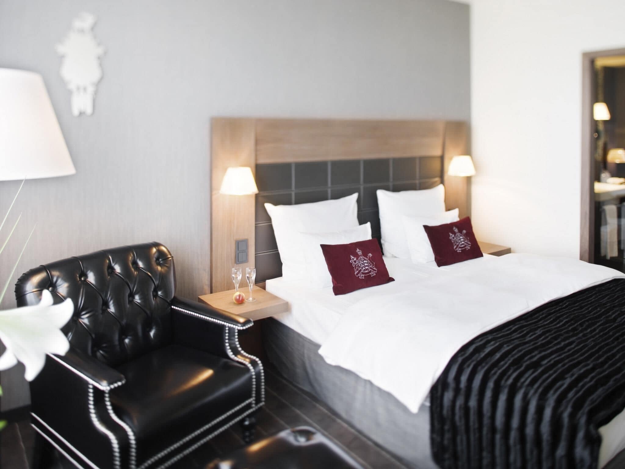Una habitación de hotel con una cama, un sillón de cuero negro, una mesita de noche con dos vasos y una lámpara de pared.