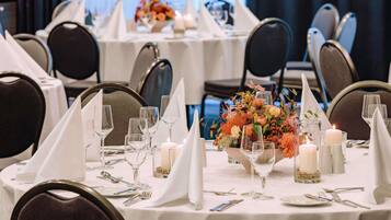 Uma sala de banquetes com mesas redondas dispostas para refeições, cada uma com toalhas brancas, copos e velas.