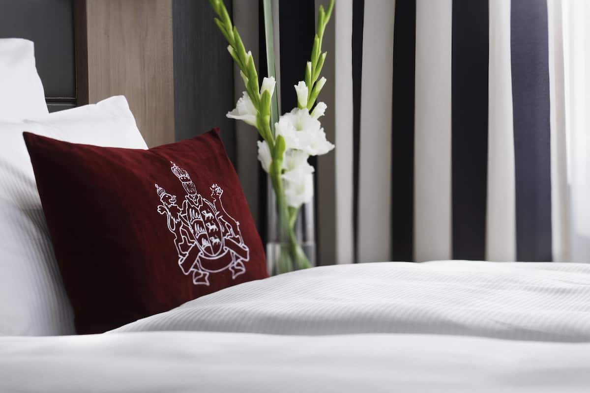 A neatly made bed with white linens, a dark red pillow with a crest design, and a vase with white flowers.