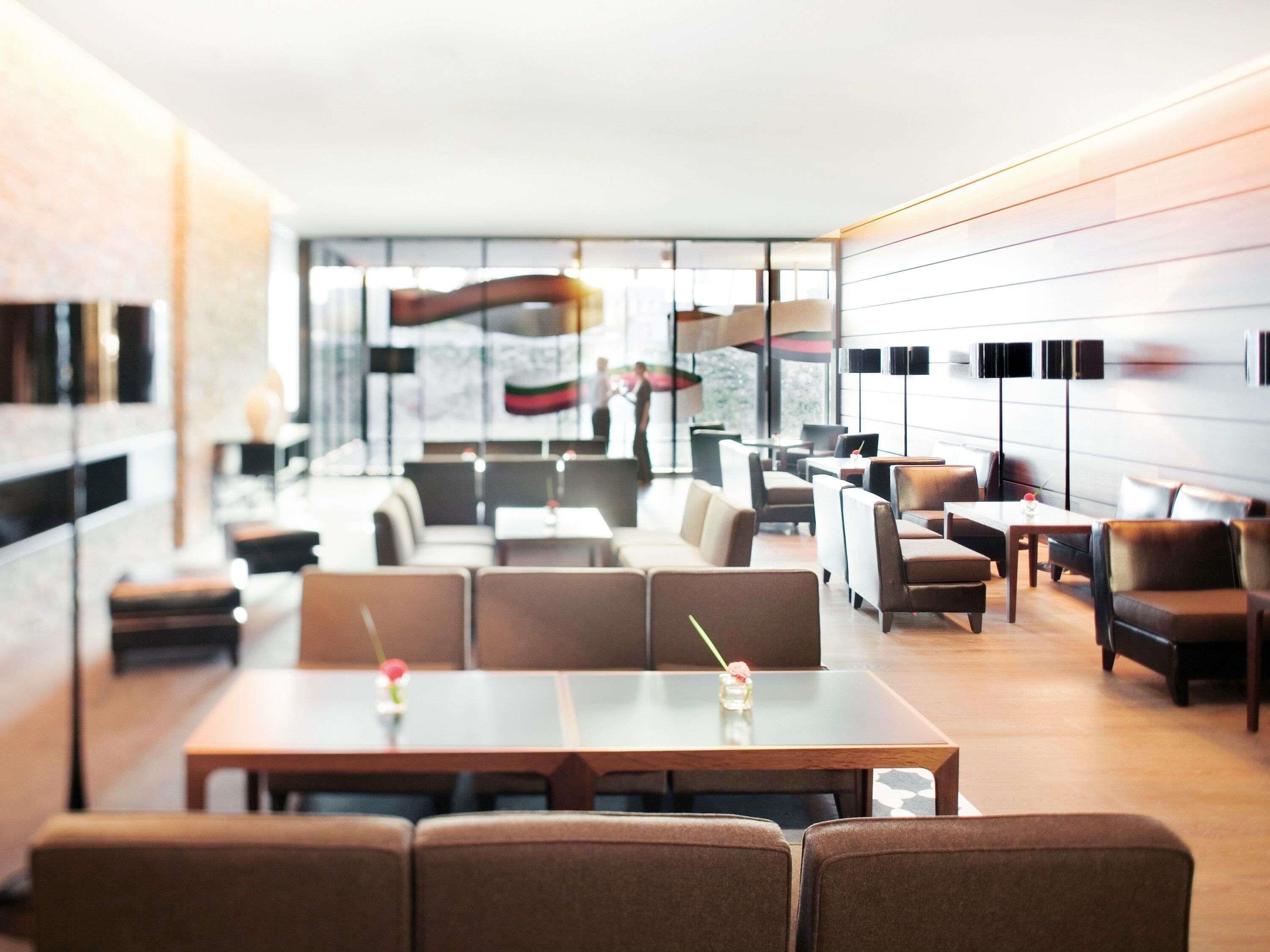 A modern hotel lobby with a central seating area, a bar, and a large window offering a view of the outdoors.
